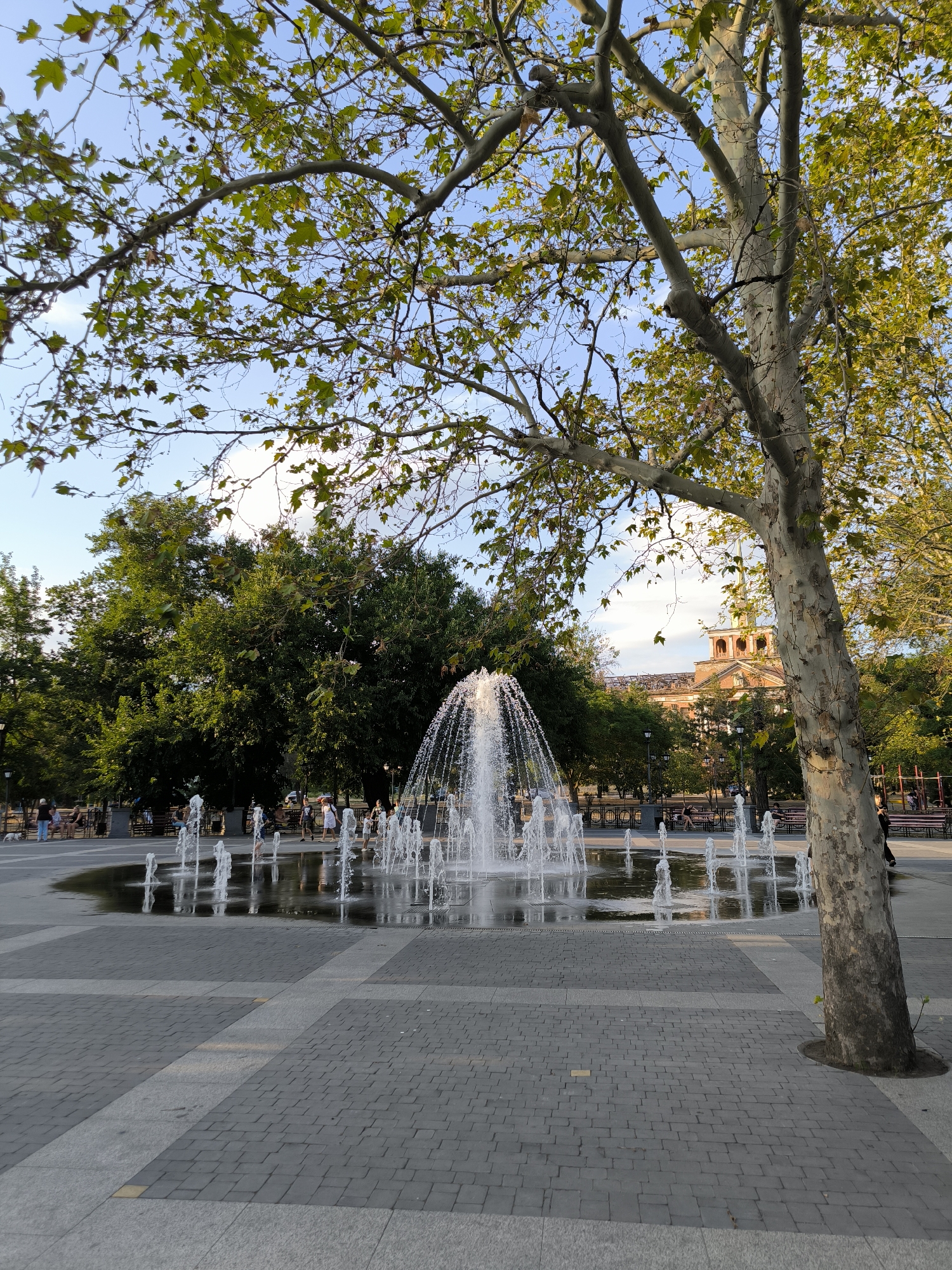 The image depicts an outdoor public space with a fountain in the foreground. The fountain has multiple jets of water spraying upwards, creating a dynamic visual effect. The area surrounding the fountain is paved with interlocking stone tiles, with a patterned arrangement. A large tree with a textured bark is prominently featured on the right side of the frame, its branches extending over the scene.
In the background, there are lush green trees, suggesting a park-like setting. Beyond the trees, a building with a distinctive architectural style, featuring arches and a tower, is visible. The sky is blue with scattered clouds.
Several people are present in the scene, some seated on benches and others standing. Some appear to be enjoying the fountain and the outdoor environment. The overall atmosphere suggests a pleasant daytime gathering in a city park.
The specific location in the city of Mykolaiv, Ukraine, is likely the **Central City Park (also known as Victory Park)**, a well-known public area that features a large fountain and is popular for recreation. The architectural style of the building in the background could potentially be identified as a landmark within or near the park, further confirming this location.