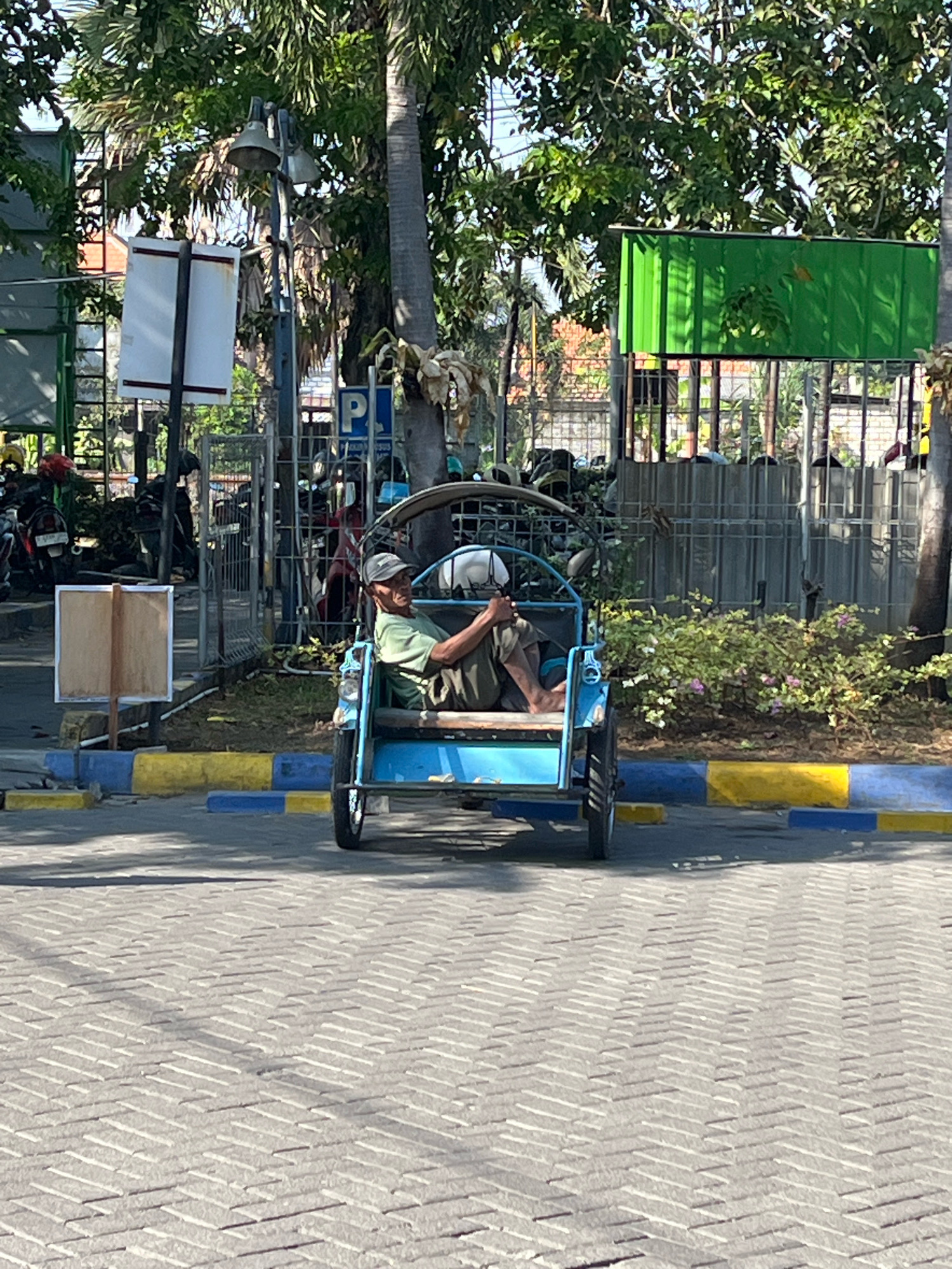 An elderly man in a green shirt and a baseball cap sits in the driver's seat of a blue motorized rickshaw, facing forward. He is holding the steering mechanism with both hands. The rickshaw is parked on a paved area marked with yellow and blue lines. In the background, a metal fence encloses an area with parked motorcycles and some greenery. Behind the fence, a bright green corrugated metal structure is visible, along with trees and buildings. The scene appears to be outdoors in a public space, possibly a parking area or street in Dapur, Indonesia, given the architecture and vegetation.