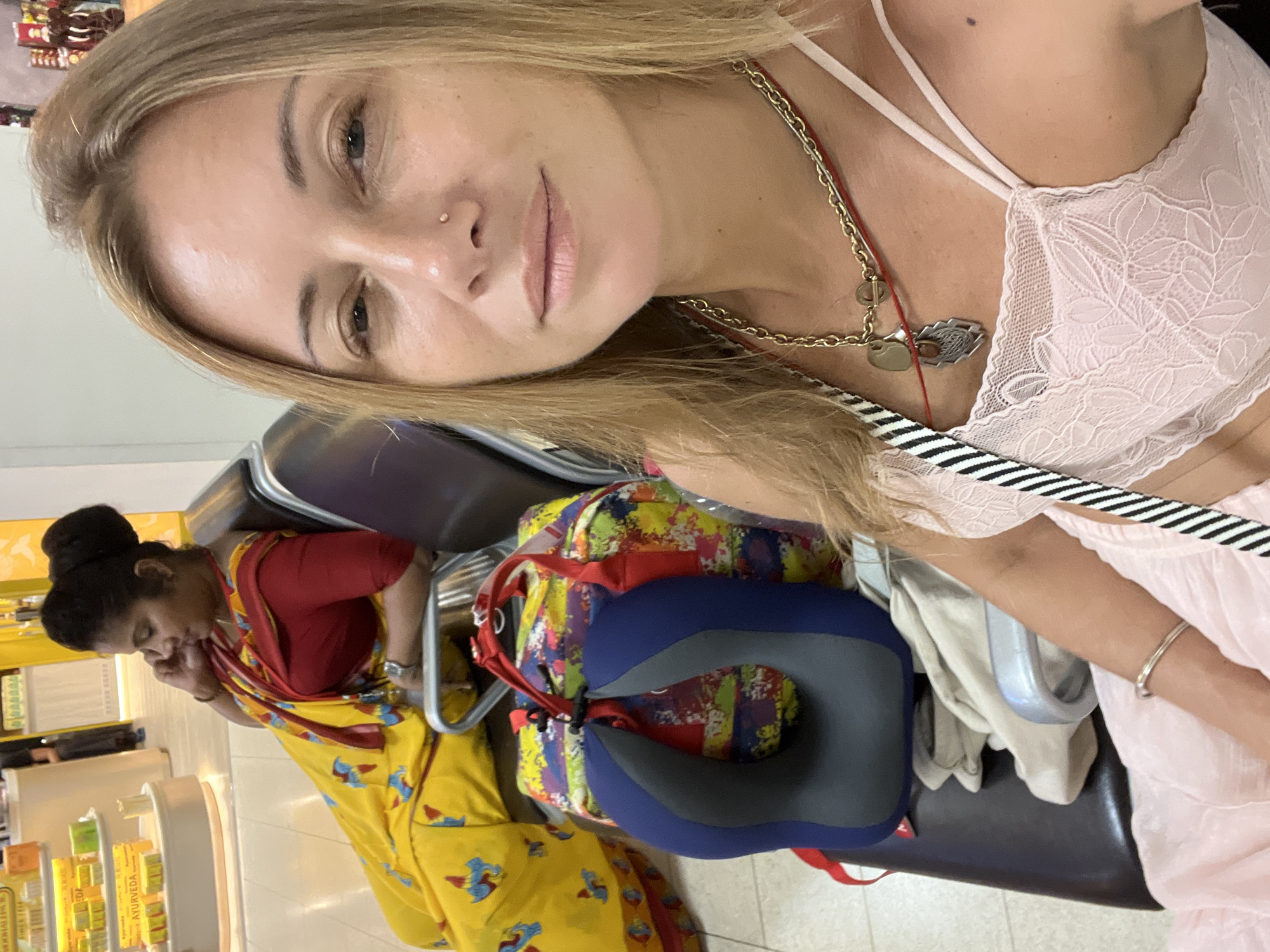 In Katunayake, Sri Lanka, two women are seated in a waiting area, likely at the airport, based on the presence of a neck pillow and luggage. In the foreground, a blonde woman is taking a selfie. She is wearing a pink lace camisole and a multi-layered necklace. Behind her, a woman dressed in a traditional yellow sari with a red blouse is seated, looking down and speaking on her phone. A colorful duffel bag and a blue neck pillow are positioned between the two women. The background shows shelves stocked with products, suggesting a retail environment.