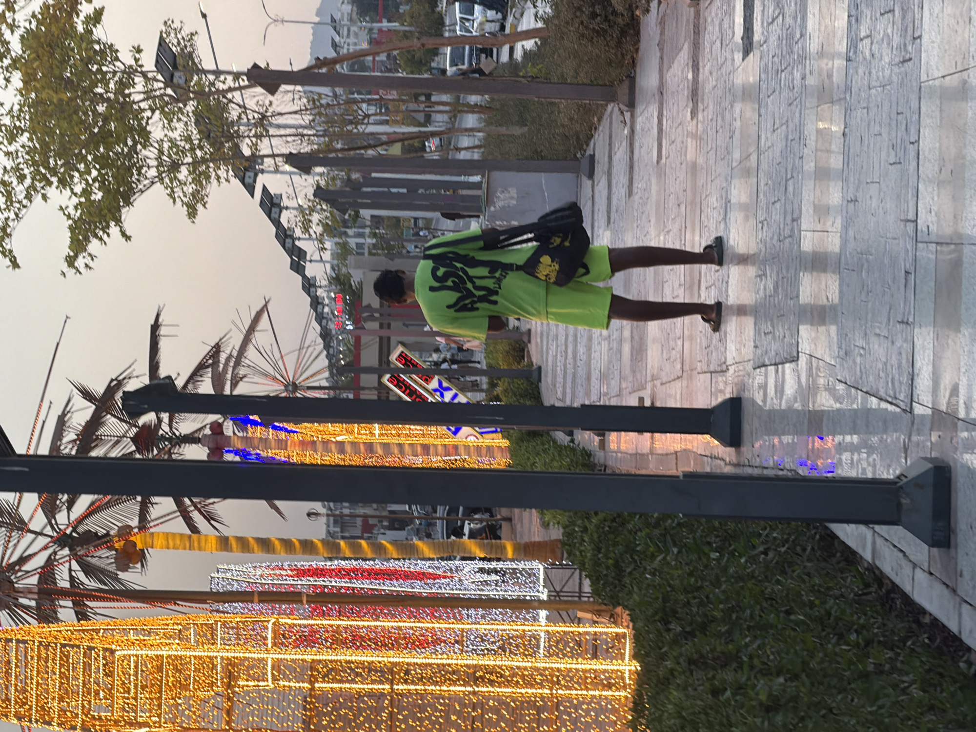 In Sharm El Sheikh, Egypt, a man stands on a paved walkway adorned with decorative lights, possibly for a festive occasion. He is wearing a bright green t-shirt with a graphic design and matching green shorts, along with dark sandals. A black bag with yellow accents hangs over his shoulder. The walkway is lined with lush green bushes and palm trees, some decorated with colorful lights. In the background, solar panels are visible along a street, suggesting a modern infrastructure. Cars and other pedestrians are present in the distance, indicating an urban setting. The overall scene suggests a lively and well-lit public space.