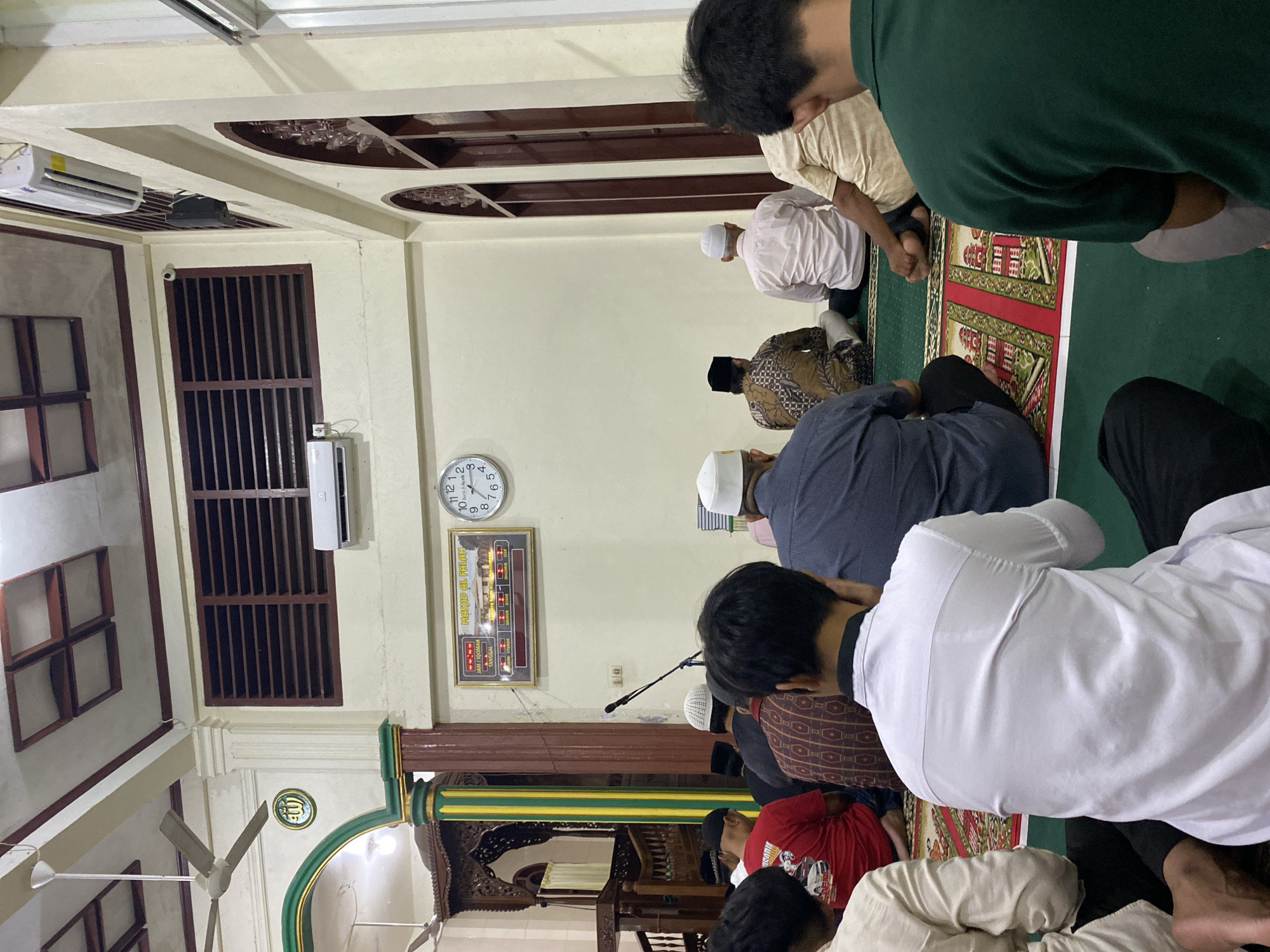 The image depicts the interior of the Masjid Al-Falah in Lhokseumawe, Indonesia, during a prayer service. Several men are seated on prayer mats arranged on a green carpeted floor. The men are facing the front of the mosque, where a minbar (pulpit) is visible on the left. The mosque's interior features cream-colored walls, a high ceiling with wooden accents, and large, louvered windows. An air conditioning unit is mounted on the wall above the windows. A digital clock displaying prayer times is also visible on the wall, along with a circular analog clock. The lighting appears to be artificial, creating a warm ambiance. The scene suggests a moment of communal prayer and reflection within the religious space.