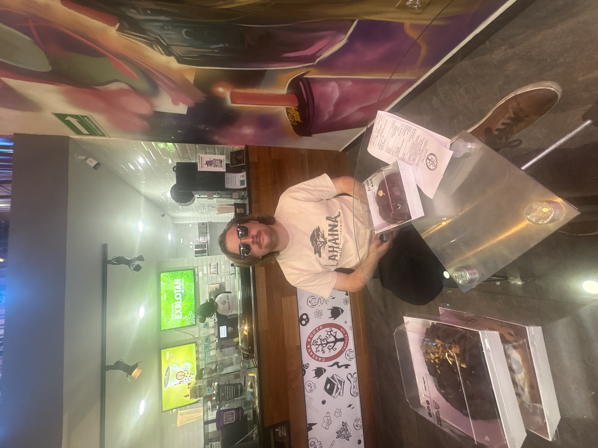 Here is a forensic description of the image:
The image shows an individual sitting at a glass table inside what appears to be a dessert shop in Huixquilucan de Degollado, Mexico. The individual is wearing sunglasses and a t-shirt with "Lahaina" printed on it. The interior is modern, with decorative art on the walls and what appear to be menus or promotional displays on the storefront windows. A long decorative banner with the words "Cassava Roots Tapioca Tea" written on it is also visible. On the table, there is a box with a chocolate dessert inside, and an order receipt. A pair of brown sneakers is positioned near the edge of the table. The scene suggests a casual setting, possibly a customer enjoying their purchase at the establishment.