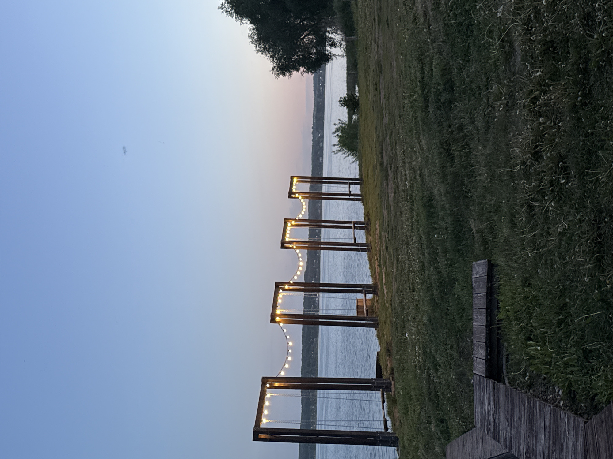 The image shows a line of swing sets overlooking a body of water, likely a lake or wide river, at dusk. The swings are dark wood frames with chains suspending the seats. String lights are strung across the tops of the swing frames. The sky fades from blue to pink towards the horizon, suggesting the sun has recently set. The opposite shore of the water is visible in the distance with trees and some hints of buildings. The swing sets are situated on a grassy area. There are boards next to the grass, possibly part of a dock or walkway.
While Votkinsk, Russia, has a large artificial lake, without additional contextual information, it cannot be definitively confirmed that this image was taken there.