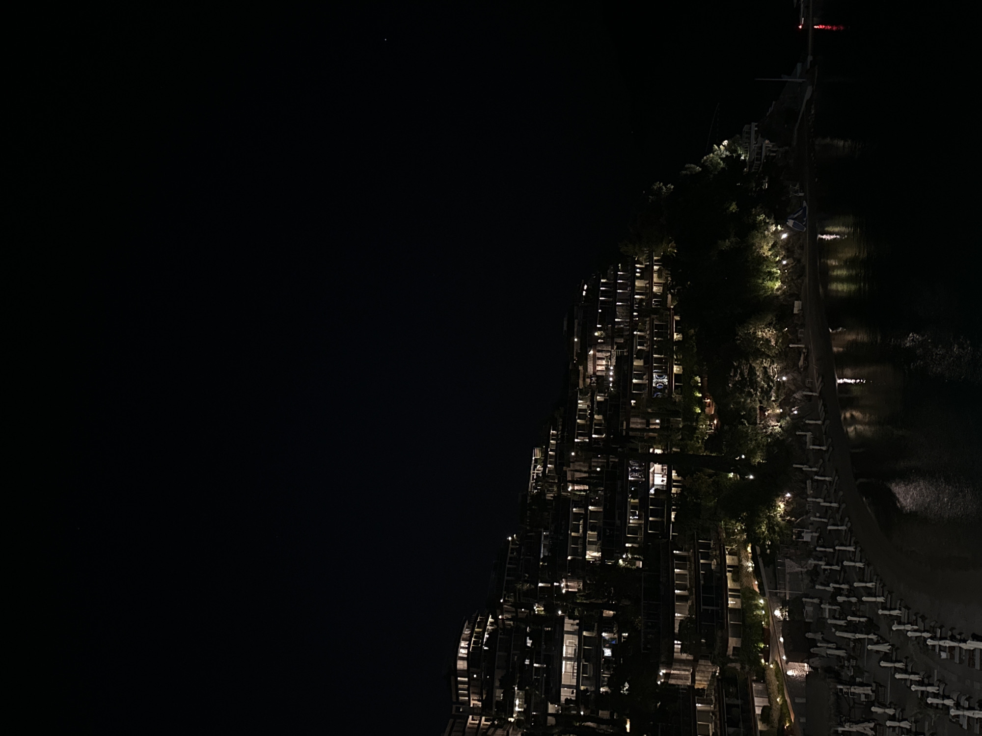 The image shows a nighttime view of a seaside resort in Budva, Montenegro. A tall building, likely a hotel or apartment complex, is illuminated with lights. Balconies and windows are visible, some with lights on. A line of trees and foliage runs along the edge of the building. Adjacent to the building is a row of beach umbrellas lining the shoreline, also illuminated by artificial lights. The water appears dark with some light reflection. The sky is dark and almost completely black.