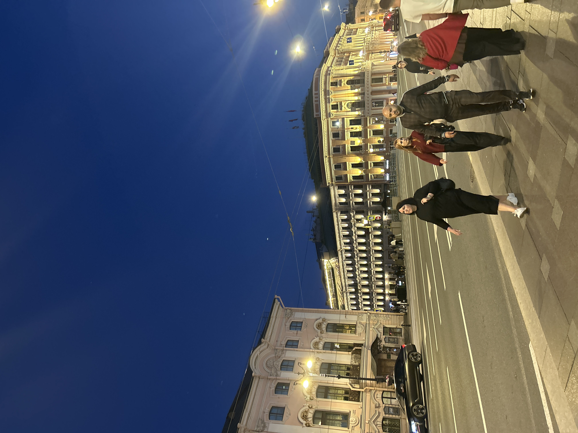In the city of Unknown, Unknown, at dusk, a street scene unfolds. The sky is a deep blue, transitioning from day to night, with a few distant lights visible. The foreground shows a sidewalk with several pedestrians. A woman in a black dress and white sneakers walks from left to right, carrying a dark bag. Behind her, a younger woman in a maroon sweater and dark pants also walks. A man in a dark patterned tracksuit and dark pants is in the center, walking towards the viewer. To his right, a woman in a red shawl over a dark outfit is walking. Another person, partially obscured, is visible behind her. A man in a white t-shirt and light-colored pants is at the far right, walking away from the viewer.
Across the street, a multi-lane road is visible with white lane markings. A dark-colored sedan is parked in the left lane. On the right side of the road, a red car is in motion. Several buildings line the street. The building on the left is a pale pink structure with ornate architectural details, including arched windows and decorative moldings. A street lamp with a bright yellow light stands on the sidewalk near this building.
Further down the street, a larger building with a pale facade is illuminated by numerous lights, highlighting its windows and architectural features. This building appears to have multiple stories and a grand facade. The overall impression is that of a busy urban environment at the end of the day, with people moving along the sidewalk and traffic on the road. The lighting suggests artificial lights are predominantly illuminating the scene, with the natural light fading.