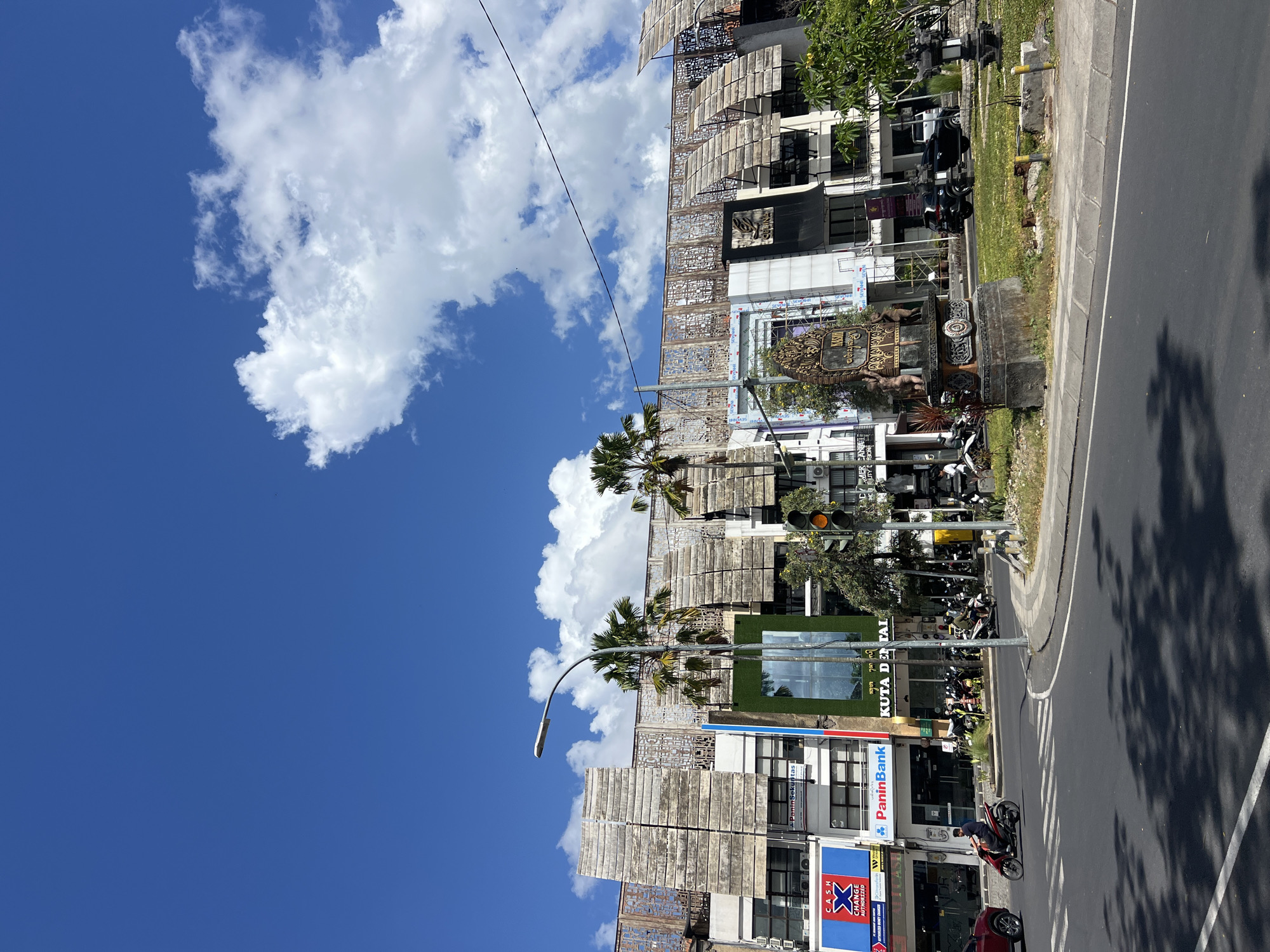 The image shows a street scene in Kuta, Indonesia, on a sunny day with a bright blue sky and scattered white clouds. The foreground features a road with traffic, including a red car on the left and several motorcycles. A person is riding a motorcycle on the road, and there are parked motorcycles on the side. The middle ground shows a building with a unique architectural design, featuring horizontal wooden louvers and decorative screens. Several businesses are visible on the ground floor of the building, including "CASH CHANGE AUTHORIZED," "PaninBank," and "KUTA DENTAL." Palm trees are planted in front of the building. A traffic light is visible on the right side of the image. The background is dominated by the sky. In the center of the image, a statue is located on a grassy area between the road and the building.