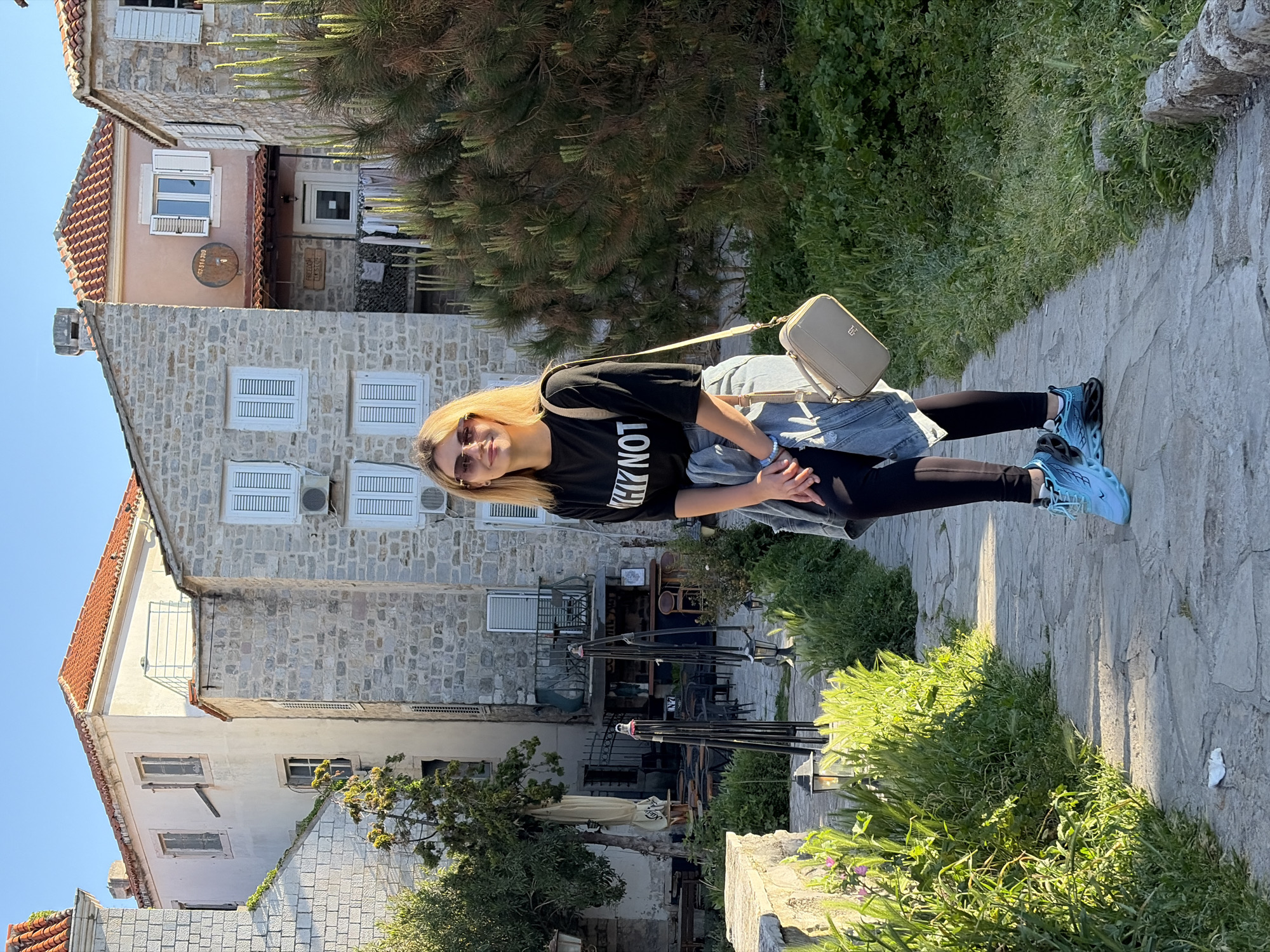 A woman stands on a stone-paved path in the city of Budva, Montenegro, posing for a photo with traditional European architecture. She is wearing sunglasses, a black t-shirt with white lettering, a denim jacket, black leggings, and light blue sneakers. She is carrying a light tan purse over her shoulder. The path is lined with greenery and leads to buildings with stone walls and red-tiled roofs. There is also a tree, outdoor seating with sun umbrellas, and a clear blue sky above.