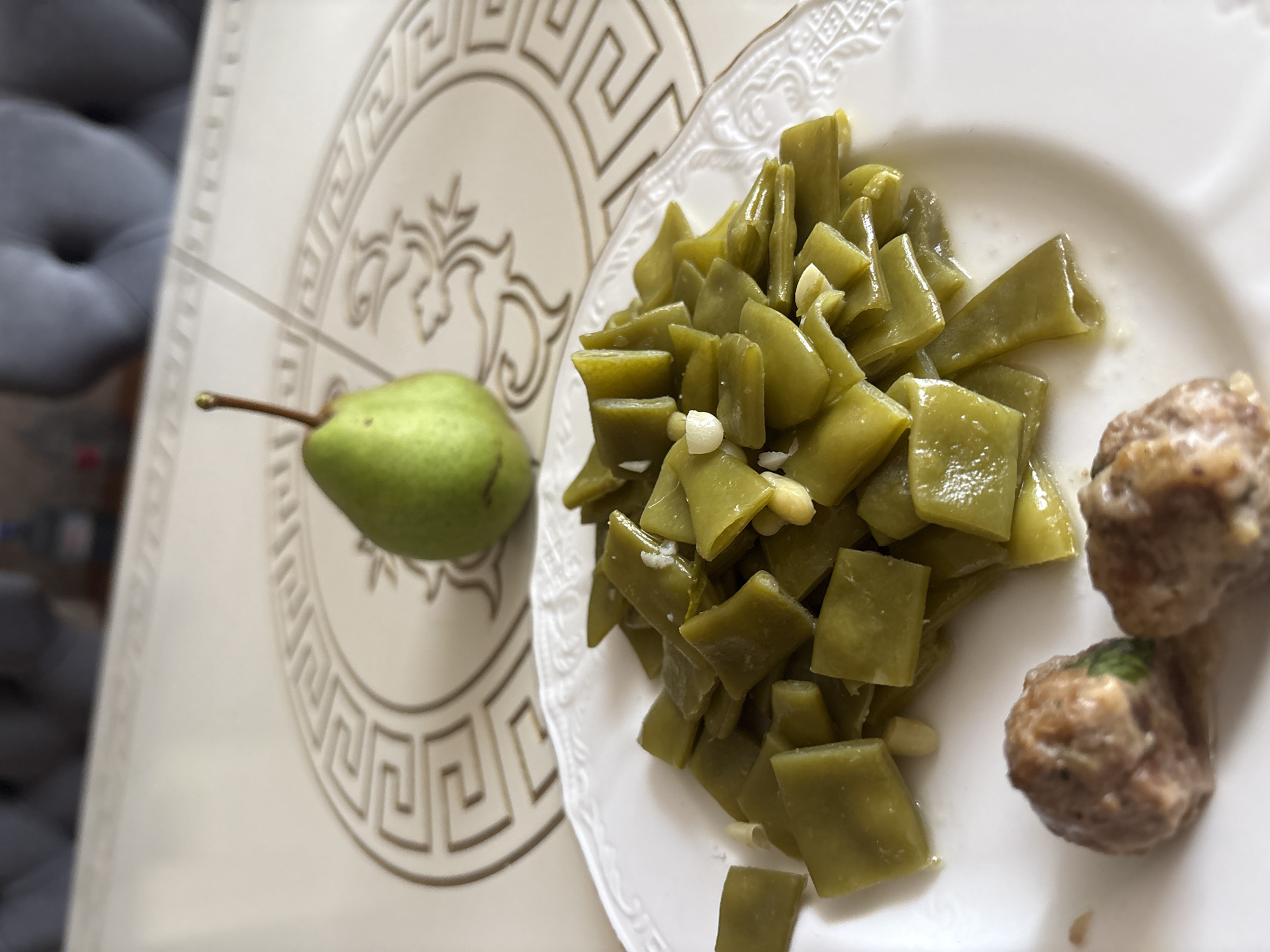 A close-up, eye-level shot shows a white plate filled with chopped green beans and two meatballs. The green beans are cut into diamond shapes and appear to be cooked with garlic. The meatballs are small and round, with a slightly browned exterior. In the background, a green pear sits on a table with a decorative pattern. The table is set against a blurred backdrop of upholstered furniture, suggesting an indoor dining setting. There are no discernible architectural or environmental features that would place this scene within the city of Georgiyevsk, Russia. The lighting is soft and natural, casting subtle shadows. The image is static and there is no indication of movement or activity.