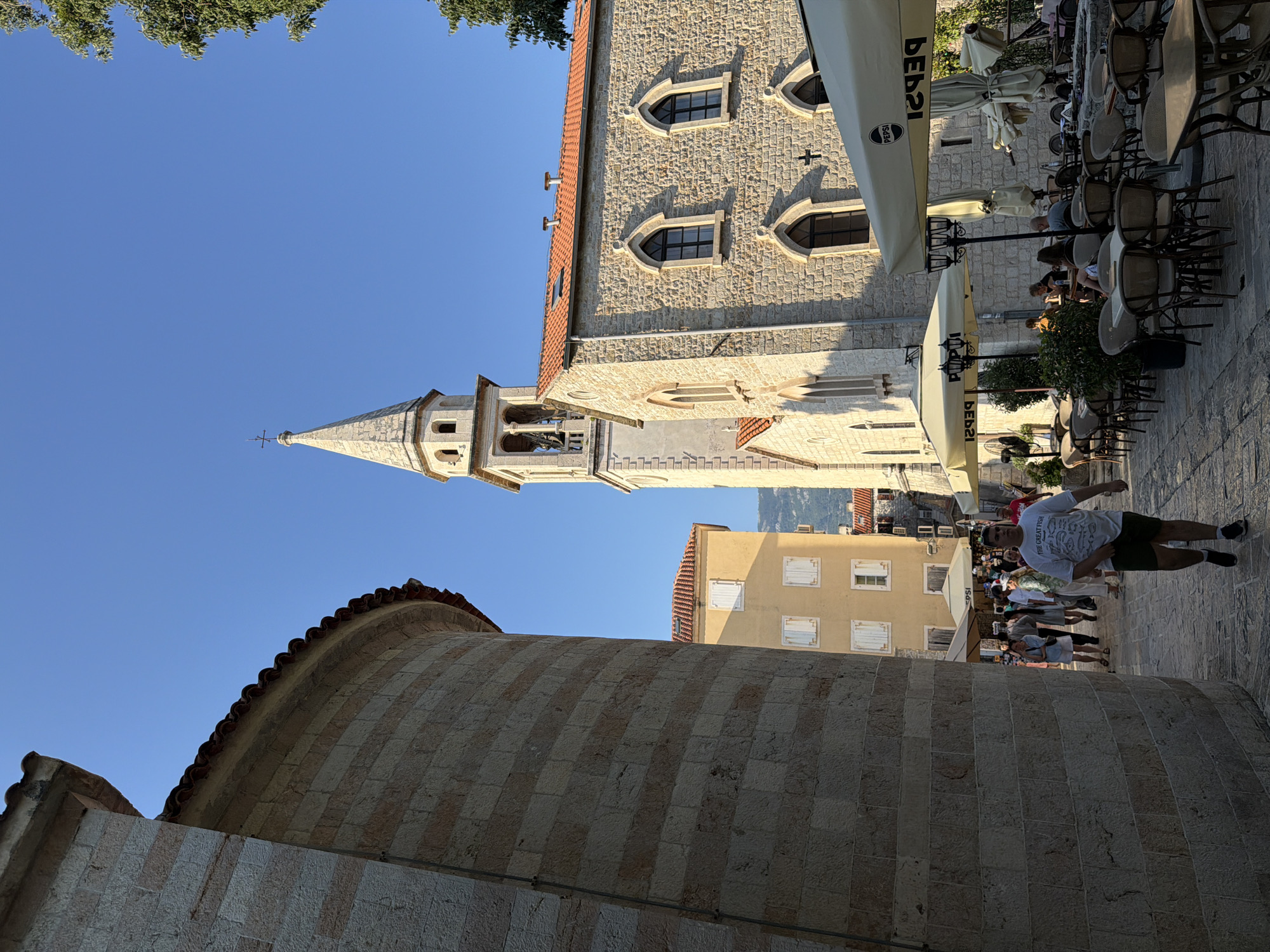 The image captures a sunlit street scene in the old town of Budva, Montenegro. On the left, a large, curved stone structure, likely part of an ancient building, dominates the foreground, casting a shadow over the cobblestone street. In the mid-ground, a bell tower with a pointed spire rises above a stone church, its facade featuring arched windows. To the right of the church, outdoor seating for a cafe is arranged under umbrellas, with patrons seated at tables. The street is bustling with activity; several individuals are walking, some appearing to be tourists, while others are seated at the cafe. The overall ambiance suggests a warm day, with a clear blue sky overhead and sunlight illuminating the historic architecture. The scene is set against a backdrop of other buildings and distant hills.