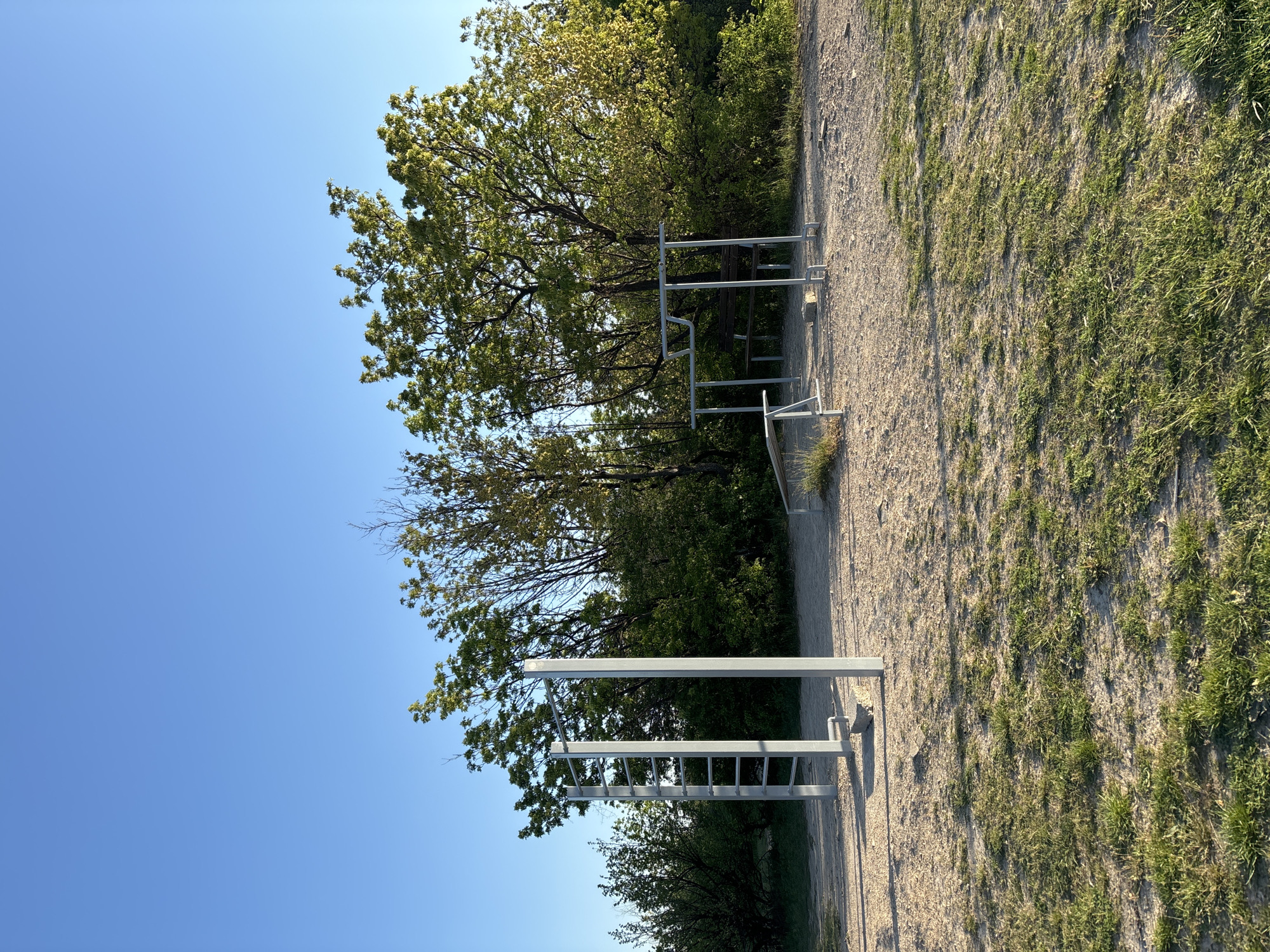 The image shows an outdoor exercise area with metal structures. The area has a gravel path adjacent to grassy terrain. The sky is clear and blue, indicating a sunny day. The location appears to be in a natural area with trees surrounding the exercise equipment. There are no visible people.