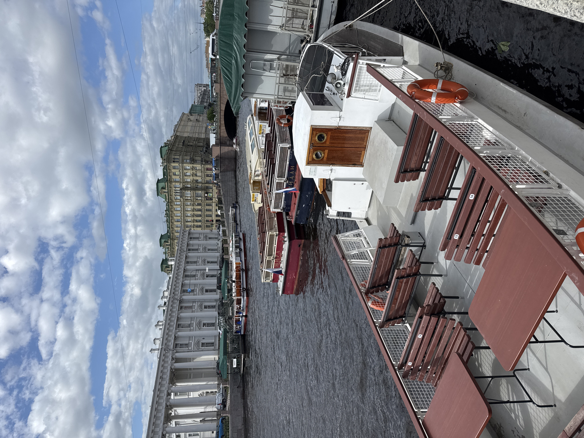 The image depicts a canal scene in Saint Petersburg, Russia. Several tourist boats are docked along a stone embankment. The boats are of various sizes, with one large white boat in the foreground featuring brown wooden seating. The water is dark and reflects the buildings lining the canal. On the left, a large white building with prominent columns is visible. Beyond it, other buildings in various architectural styles stretch along the canal. The sky is partly cloudy. The scene is captured in daylight.