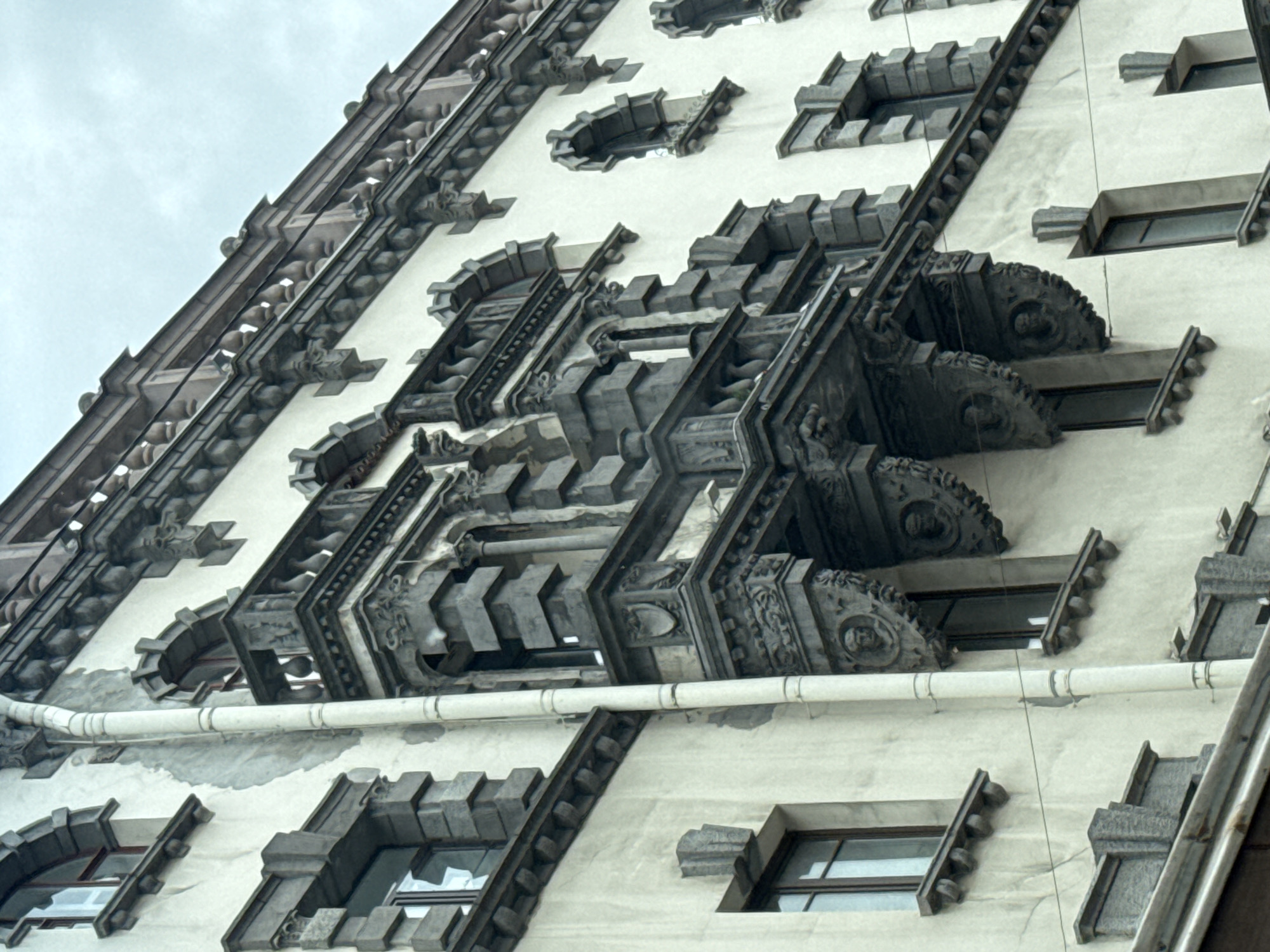 This media file is a close-up, angled photograph of the ornate facade of a building, likely an apartment building or hotel, in Saint Petersburg, Russia. The building features intricate Gothic-inspired architectural details, including decorative stone carvings, crenellations, and protruding window elements. The facade is primarily light-colored, possibly limestone or stucco, with dark grey or black stone ornamentation.
The photograph focuses on a section of the building's upper floors, showcasing repetitive patterns of windows and decorative structures. Several windows are visible, some appearing recessed and others protruding with decorative hoods. The most striking elements are the elaborate, layered stone structures that jut out from the building, resembling stylized gargoyles or elaborate cornices with circular motifs. A horizontal white pipe runs across the building's facade, interrupting the otherwise consistent architectural design.
The sky in the upper left corner is overcast and a pale grey, suggesting either early morning, late evening, or a cloudy day. There is no visible text in the image. The overall impression is one of historical grandeur and detailed craftsmanship, common in the architecture of Saint Petersburg. The image is a detail shot, emphasizing the textures and forms of the building's exterior rather than a wider scene.