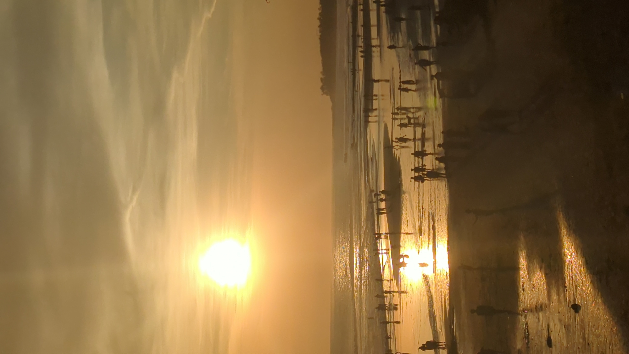 The image shows a beach at sunset, likely in North Kuta, Indonesia. The sun is setting over the water, casting a golden glow on the scene. Silhouetted figures of people are visible along the shoreline and in the shallow water, suggesting they are walking or wading. The wet sand reflects the sunlight, creating shimmering effects. The sky is filled with clouds, adding to the atmospheric quality of the sunset.