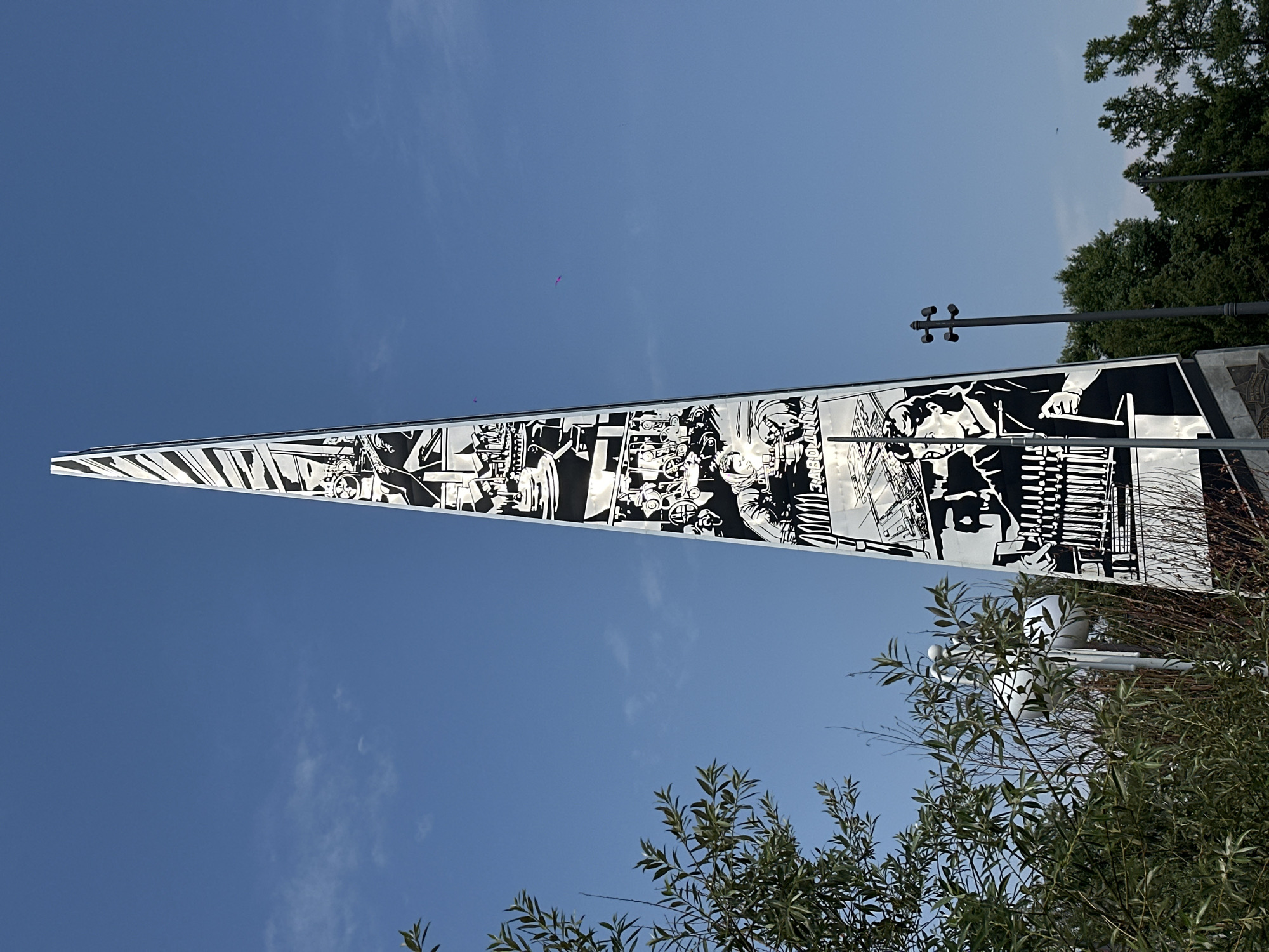 The image depicts a tall, slender obelisk monument covered in black and white graphic depictions of people and industrial scenes. The monument is situated outdoors against a clear blue sky with a few wispy clouds. To the left, lush green foliage from trees partially obscures the lower portion of the monument and extends into the foreground. To the right, a tall, dark lamppost stands near the monument, and further in the background, the tops of green trees are visible. The obelisk's surface is adorned with numerous panels, each illustrating scenes of work, possibly related to industry or manufacturing, with stylized figures and machinery. One panel clearly shows the Cyrillic inscription "ЗАВОДЛИЦ" (ZAVODLITs), which translates to "Factory." This monument is located in Barnaul, Russia, and is known as the "Monument to the Workers of the Machine-Building Plant."