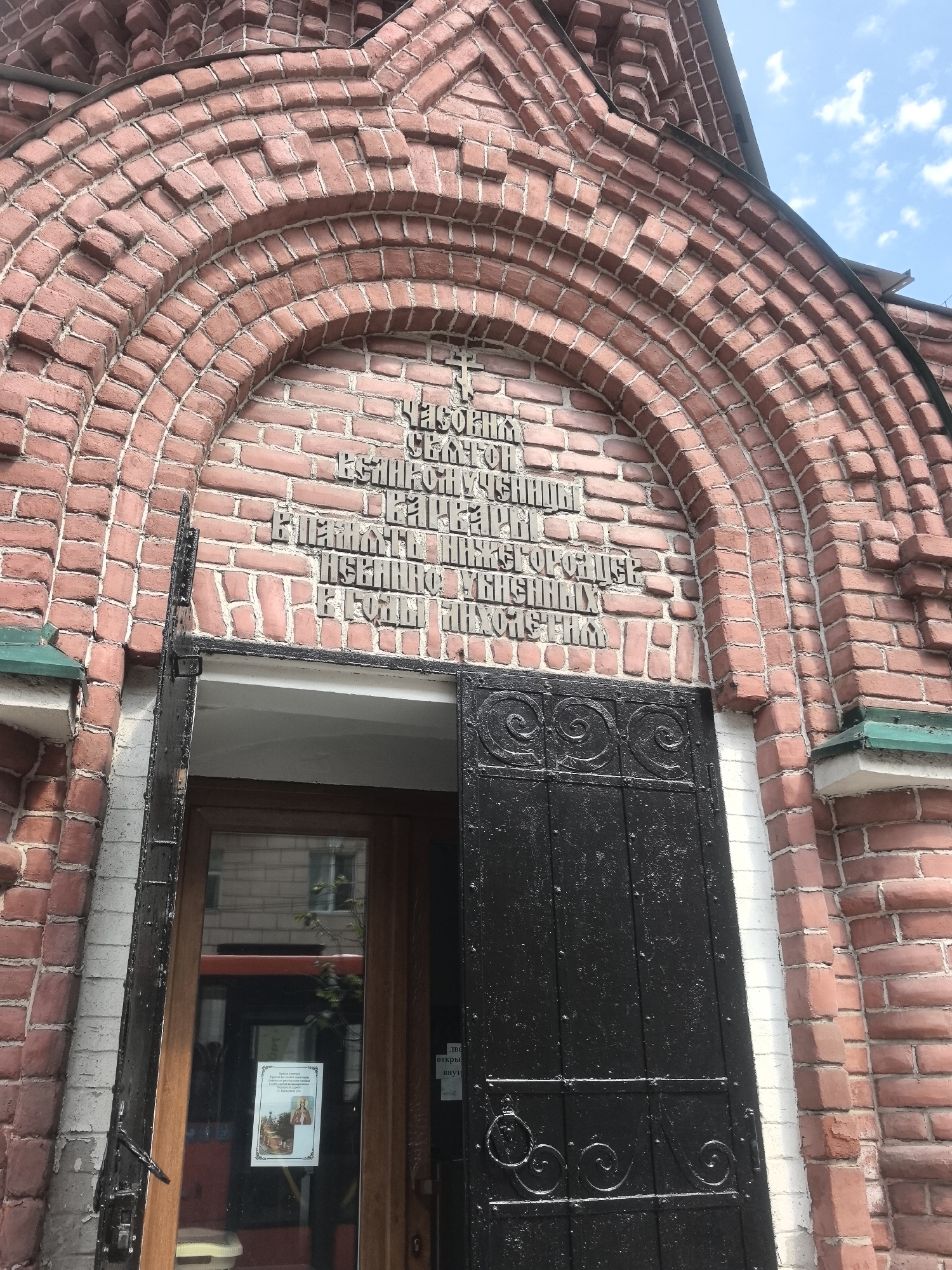 This is a low-angle, close-up, exterior shot of a brick building, likely a church, in Nizhny Novgorod, Russia. The focus is on an arched entrance with elaborate brickwork forming the arch. In the center of the arch, there is an inscription in Cyrillic lettering. Below the inscription is a large, black, wrought-iron gate. To the left of the gate is a wooden door with a glass pane. Reflected in the glass is a red bus and buildings. A white poster with an image of religious figures is affixed to the glass door. The sky is visible above the building, with some clouds. The brickwork is weathered and shows signs of age. The overall impression is one of historical architecture and religious significance.
