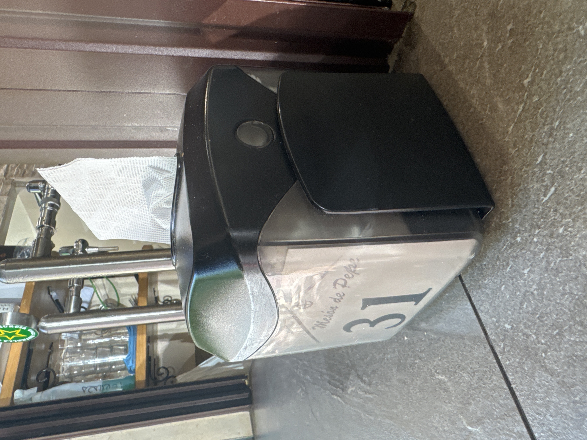 The image shows a close-up of a black napkin dispenser with a transparent compartment holding napkins. A white card with the text "Messon de Pepe" and the number "31" is visible inside the dispenser. The dispenser is placed on a grey tiled floor. In the background, there are shelves with glasses and a beer tap, suggesting a bar or restaurant setting. The overall impression is a commercial establishment. There are no people visible in the image.
Based on the text "Messon de Pepe", this establishment is likely located in Murcia, Spain, as "Messon" is a common term for a tavern or inn in Spanish cuisine. However, without more information, the exact location within Murcia cannot be determined from the image alone.