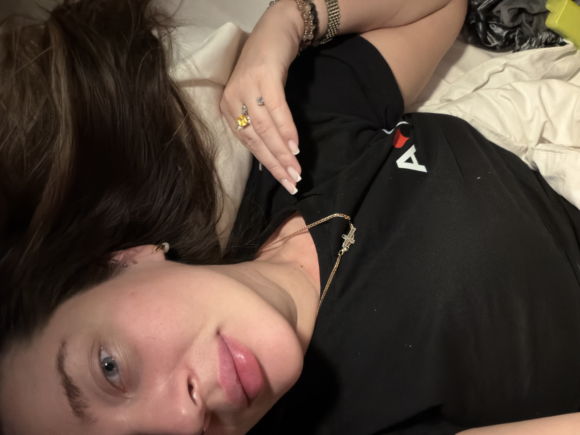 A person lies on a bed, their head resting on a pillow. They are wearing a black t-shirt, a gold necklace with a cross pendant, and multiple bracelets on their wrist. A ring with a yellow stone adorns their finger. Their hair is brown, and their eyes are blue. The bedding is predominantly white with some dark-colored fabric visible in the background. The location is not discernible from the image.