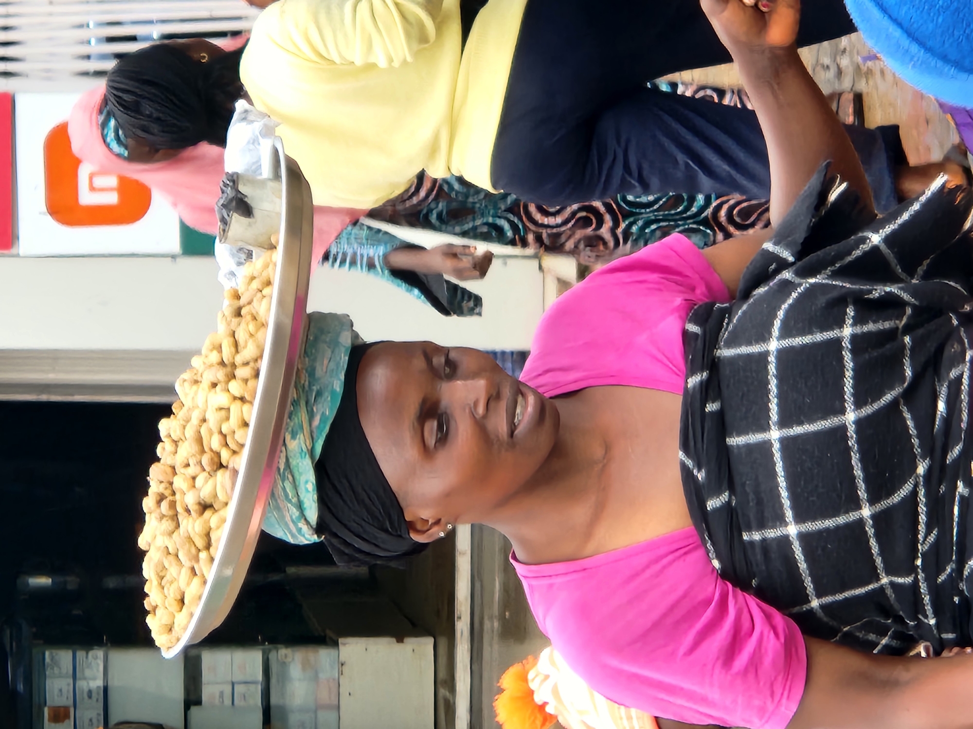 An outdoor market scene in an unknown city features a woman carrying a large metal tray of peanuts on her head. She wears a pink t-shirt and a black and white plaid wrap around her waist. To her right, another woman in a yellow shirt and dark pants stands with her back to the camera. Behind them, a woman in a pink headscarf and a patterned dress is visible. The background shows market stalls and buildings.