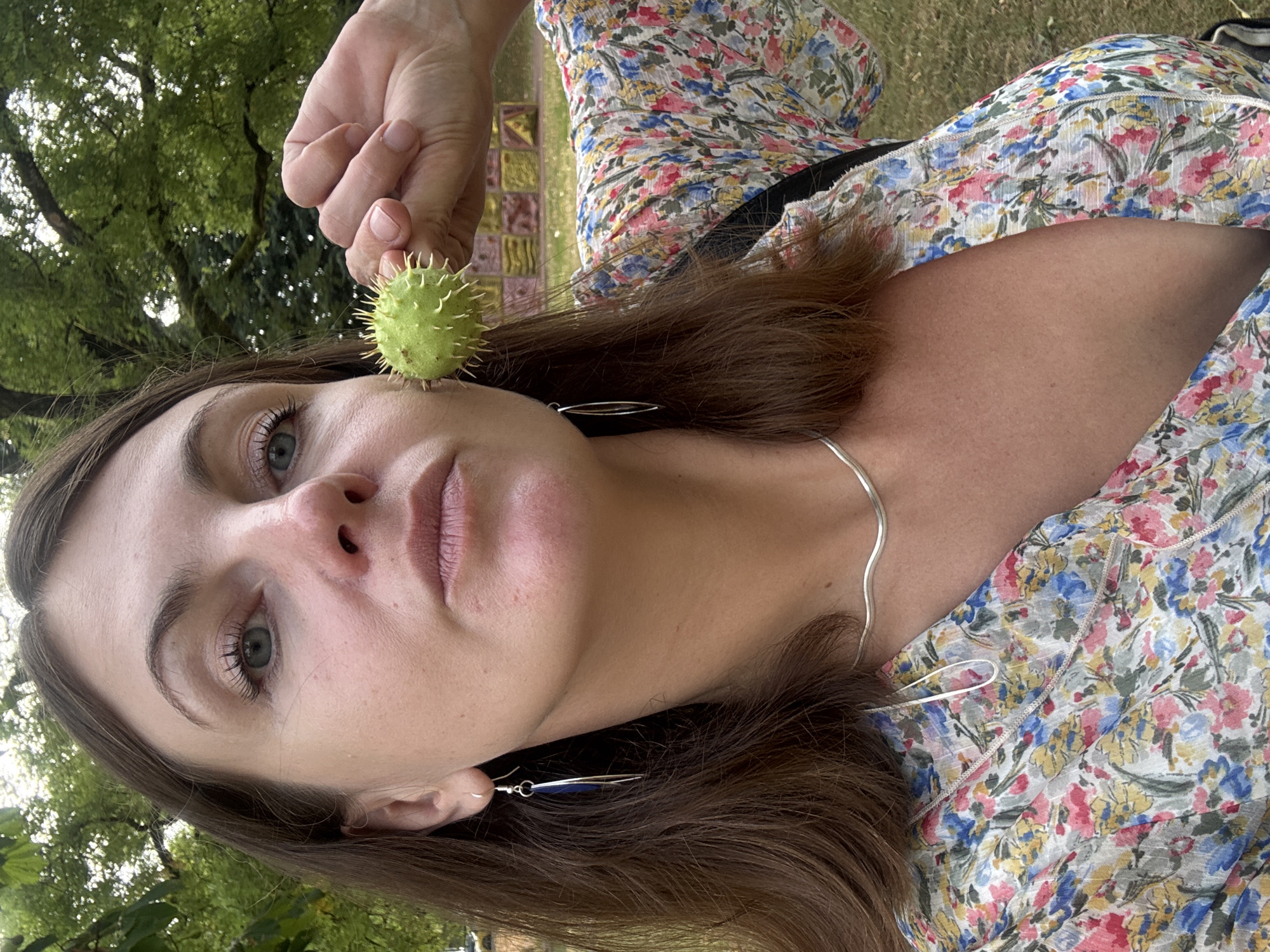 A woman is holding a spiky green fruit in front of her face outdoors, with trees and foliage in the background. She is wearing a floral-print dress and silver jewelry.