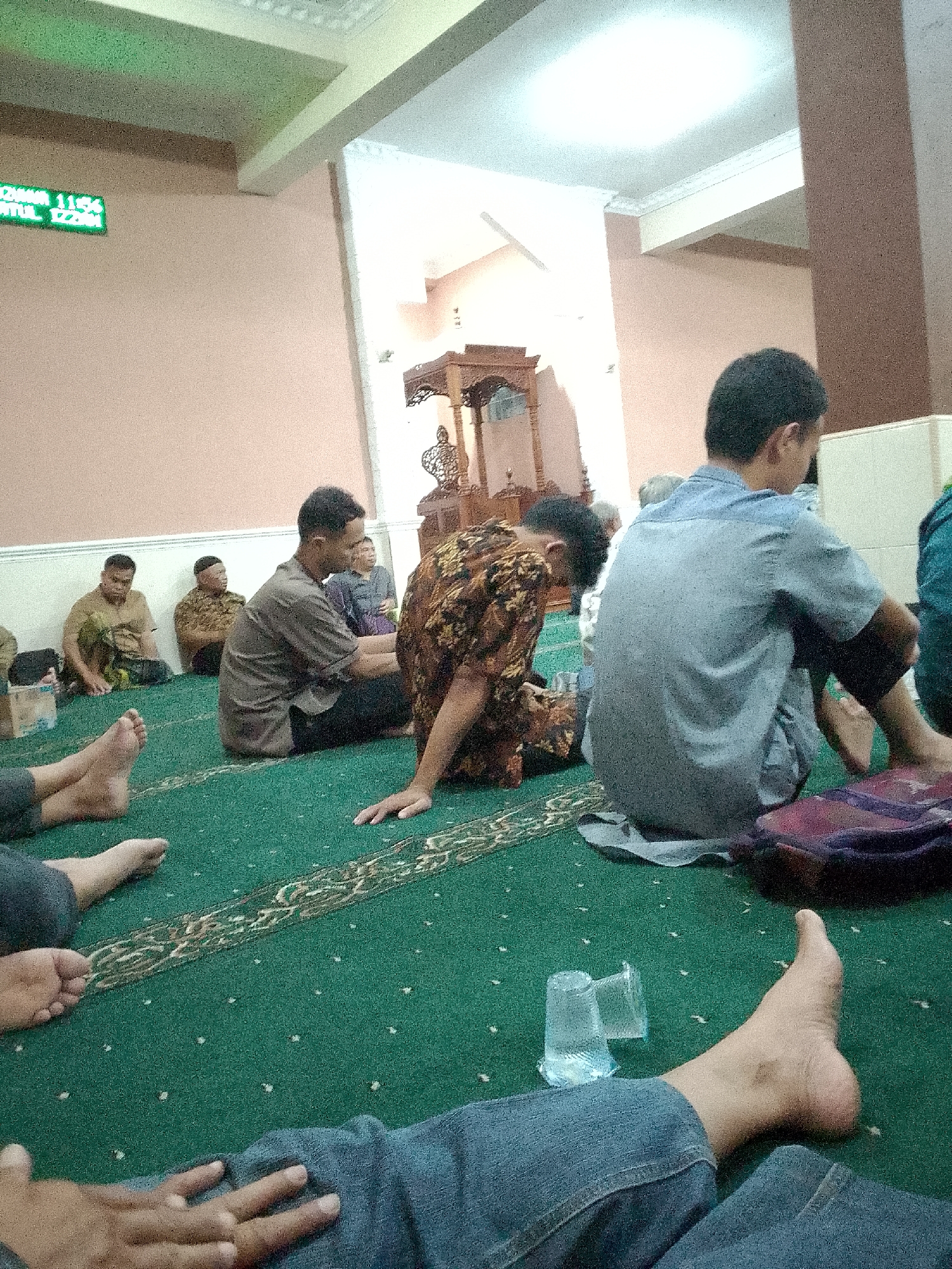 In a mosque in Kertajaya, Indonesia, several men are sitting on a green carpeted floor. The men are dressed in casual clothing, and some are facing away from the camera. One man in the foreground is wearing a blue shirt, and another to his left is wearing a patterned brown shirt. In the background, two men are seated, one in a brown shirt and another in a dark shirt. The mosque has a high ceiling with white decorative molding, and a large wooden structure that appears to be a pulpit or decorative element is visible in the background. A digital clock displaying green numbers is mounted on a light pink wall. Several plastic cups and a bag are on the floor. The overall impression is of a gathering or prayer service.