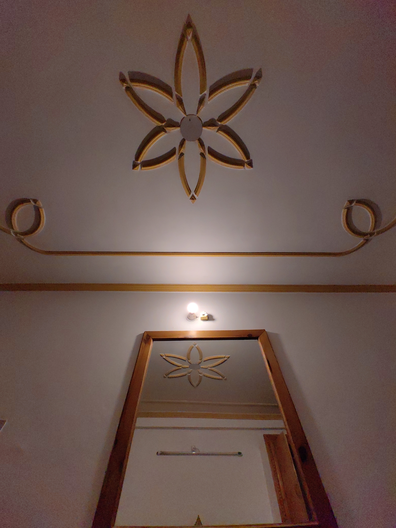 This is a low-angle, indoor shot of a ceiling and wall decorated with ornate gold trim. At the top, a symmetrical floral design is embossed into the white ceiling with gold accents. Below this, a horizontal gold line runs across the ceiling, which is flanked by two more decorative gold scrolls on either side. Further down, a single light bulb illuminates a wall. Centered on the wall is a large mirror with a wooden frame. The mirror reflects a portion of the room, including what appears to be another decorative element similar to the one on the ceiling, and a horizontal bar on the wall. The overall impression is one of decorative interior design. The image was taken in Vashisht, India.