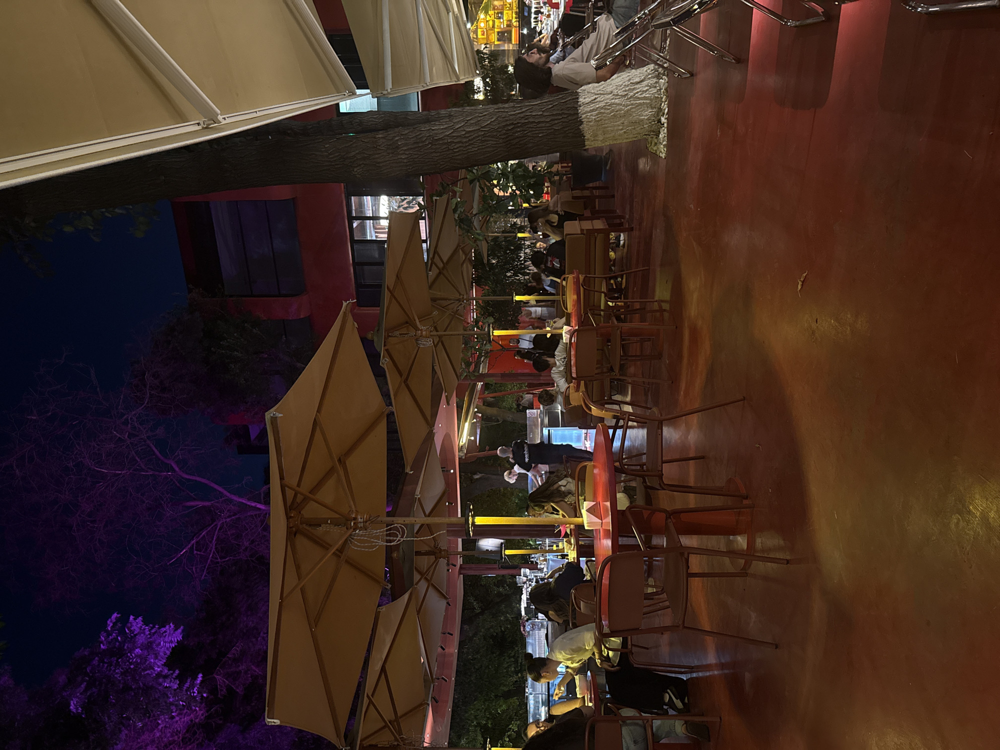 The image depicts an outdoor seating area of a restaurant or cafe at night. The area is covered by large, tan umbrellas, and the ground is a reddish-brown color, likely concrete or a similar material, reflecting ambient light. Several tables with red tops and brown chairs are arranged throughout the space. In the background, a building with red and dark sections is visible. Trees are present, some illuminated with purple lighting, creating a vibrant atmosphere. People are seated at various tables, some appearing to be dining or socializing. A server is visible behind a counter in the middle distance. The scene suggests a lively evening setting in Yerevan, Armenia.