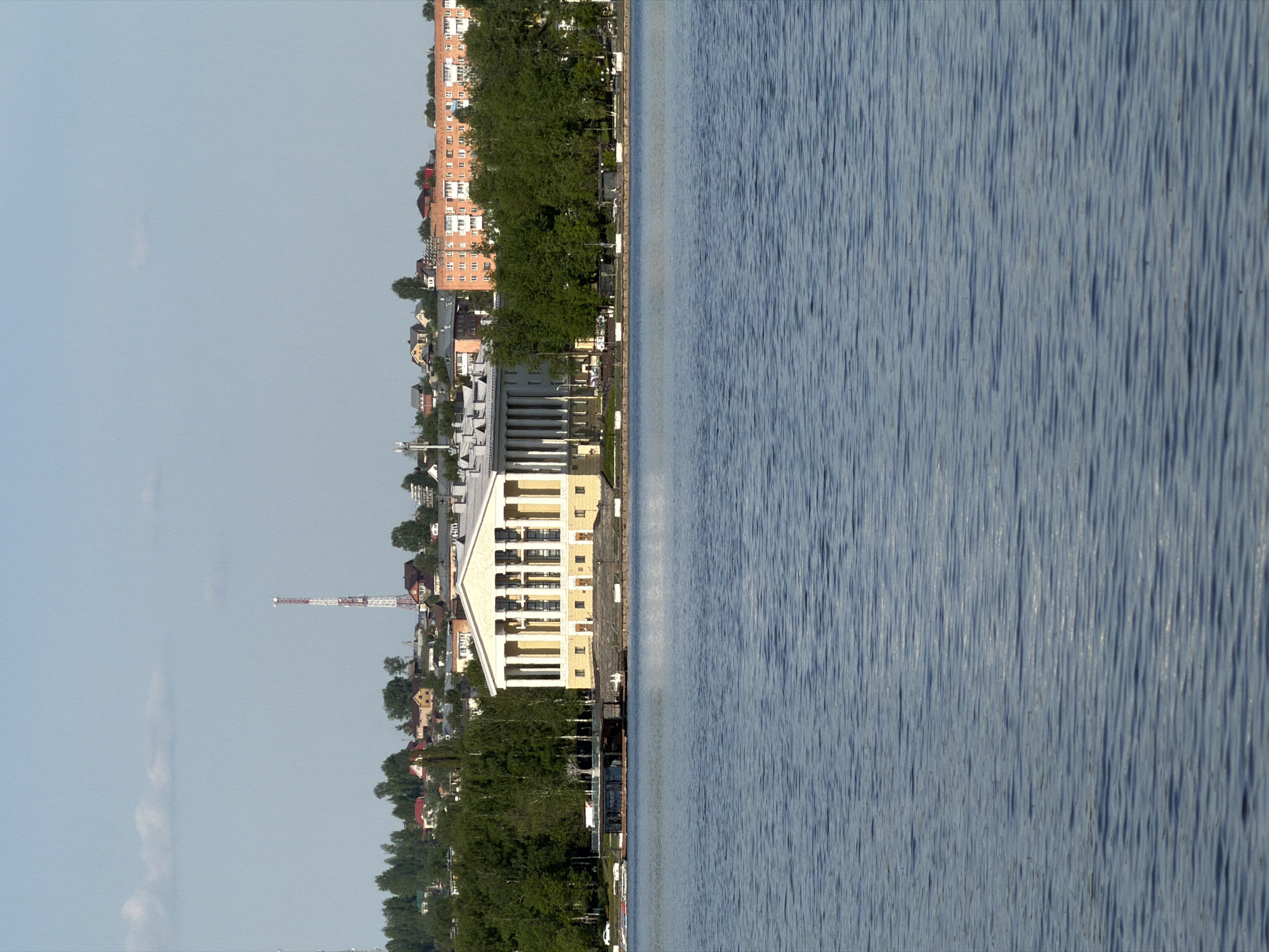The image depicts a waterfront scene in Votkinsk, Russia, viewed from the water. The right side of the frame is dominated by the blue rippled surface of the Votkinsk reservoir. The shoreline features a combination of buildings and trees. A modern, light-yellow building with white columns is prominently located along the shore, near what appears to be a paved promenade. Further inland are residential buildings, including a multi-story brick apartment building and smaller homes. A red and white crane is visible in the distance. The sky is a pale blue with some clouds.