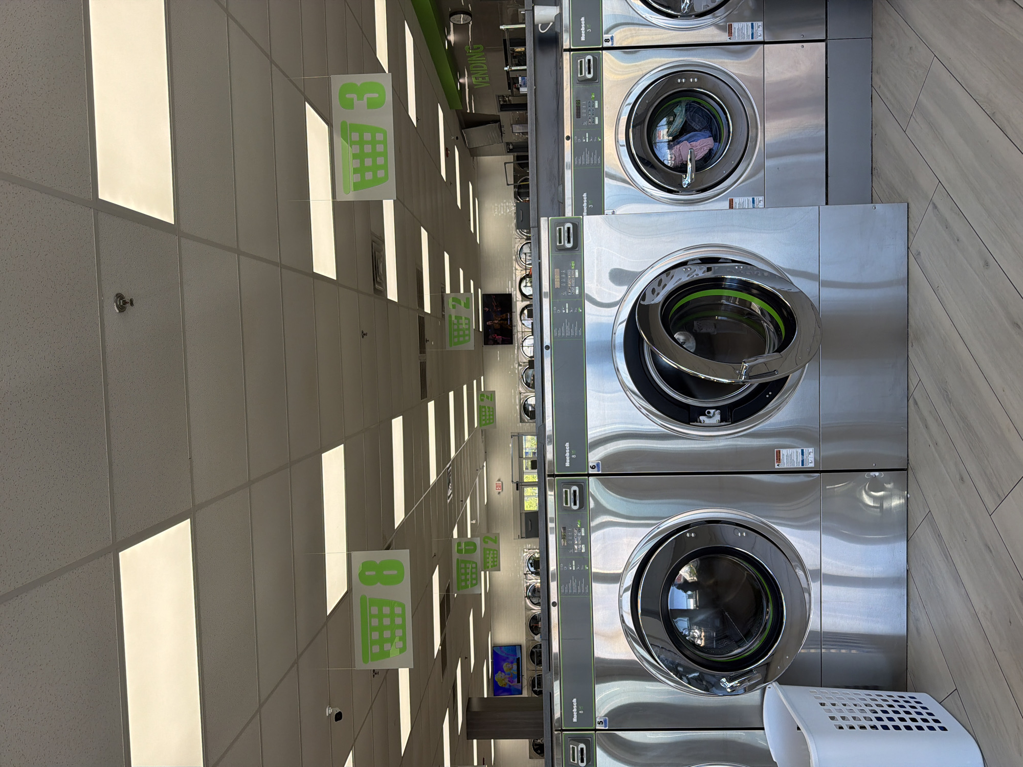 The image depicts the interior of a laundromat in Niles, United States of America. Rows of industrial-sized washing machines are stacked on top of each other on the right side of the frame. The machines are stainless steel, with some doors open and others closed, revealing some laundry inside. The left side of the frame features a white, tiled ceiling with fluorescent lighting. Hanging from the ceiling are green signs indicating the number of the corresponding washing machine row. Further down the laundromat, a television screen can be seen. A white laundry basket sits on the floor in the lower right corner of the image. The floor is tiled with light-colored wood-effect planks.