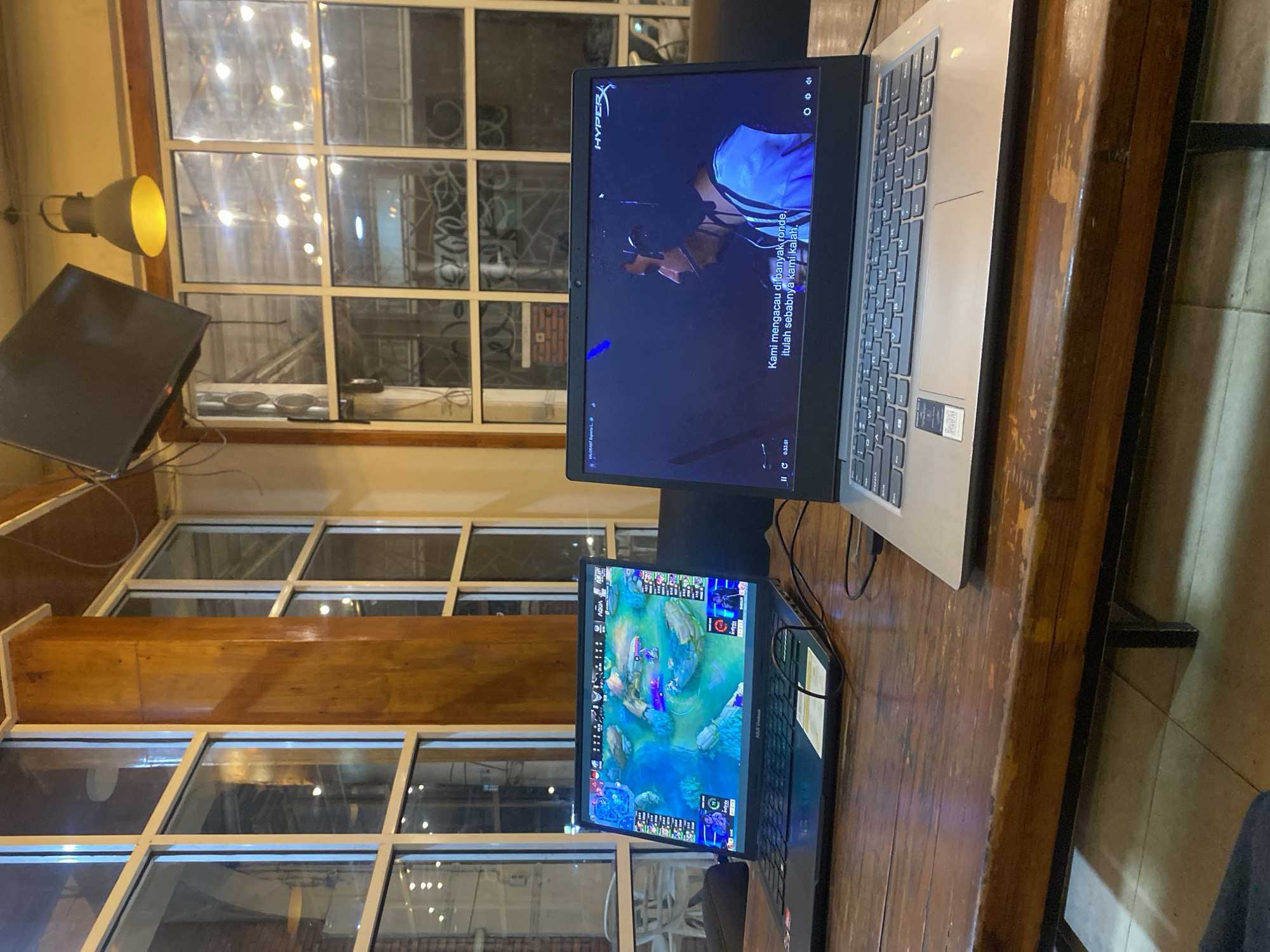 Two laptops are placed on a wooden table in what appears to be an indoor establishment with large windows. The laptop on the left displays a colorful video game interface, likely a MOBA or similar strategy game, with detailed graphics of terrain, characters, and interface elements. The laptop on the right shows a video playing, featuring a close-up of a person wearing headphones, with Indonesian subtitles displayed at the bottom of the screen. The environment suggests a cafe or gaming lounge, with industrial-style lighting fixtures and window frames visible in the background, hinting at an urban setting. The overall atmosphere is casual and focused on digital entertainment.