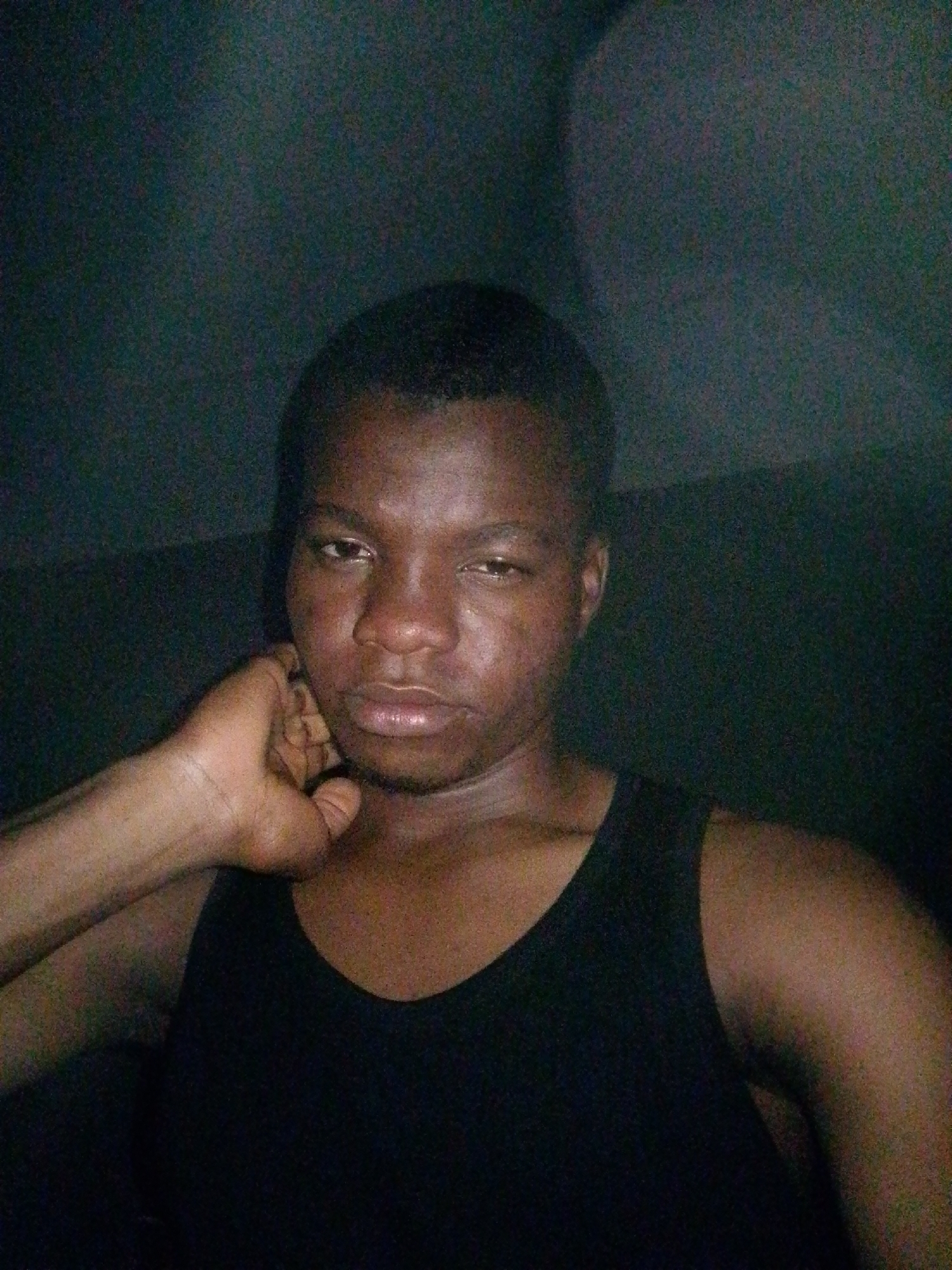 A close-up, overhead shot shows a dark-skinned young male, presumably in Ipetumodu, Nigeria, looking at the camera. The man is wearing a black sleeveless shirt. His left arm is bent and his hand is resting on his chin. The background is dark and out of focus, creating a somewhat shadowy effect. The image is slightly grainy, indicating it may have been taken in low light conditions.