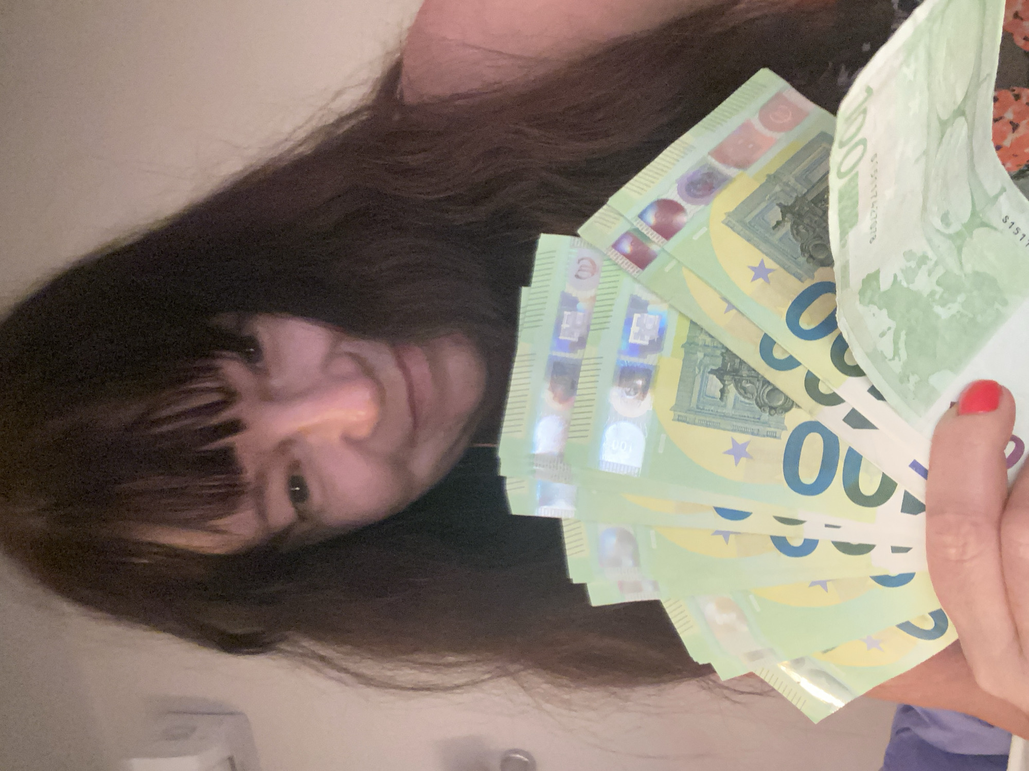 The image shows a woman holding a fan of 100 Euro banknotes. She has dark brown hair and is looking at the camera with a slight smile. The banknotes are a light green color and have the number "100" prominently displayed. The image appears to be taken indoors, with a plain wall in the background. There is no visible indication of the time of day, weather, or specific location within Novi Petrivtsi, Ukraine. The woman's expression suggests a sense of happiness or accomplishment, possibly related to the money she is holding. The banknotes have some visible text, including serial numbers and security features.