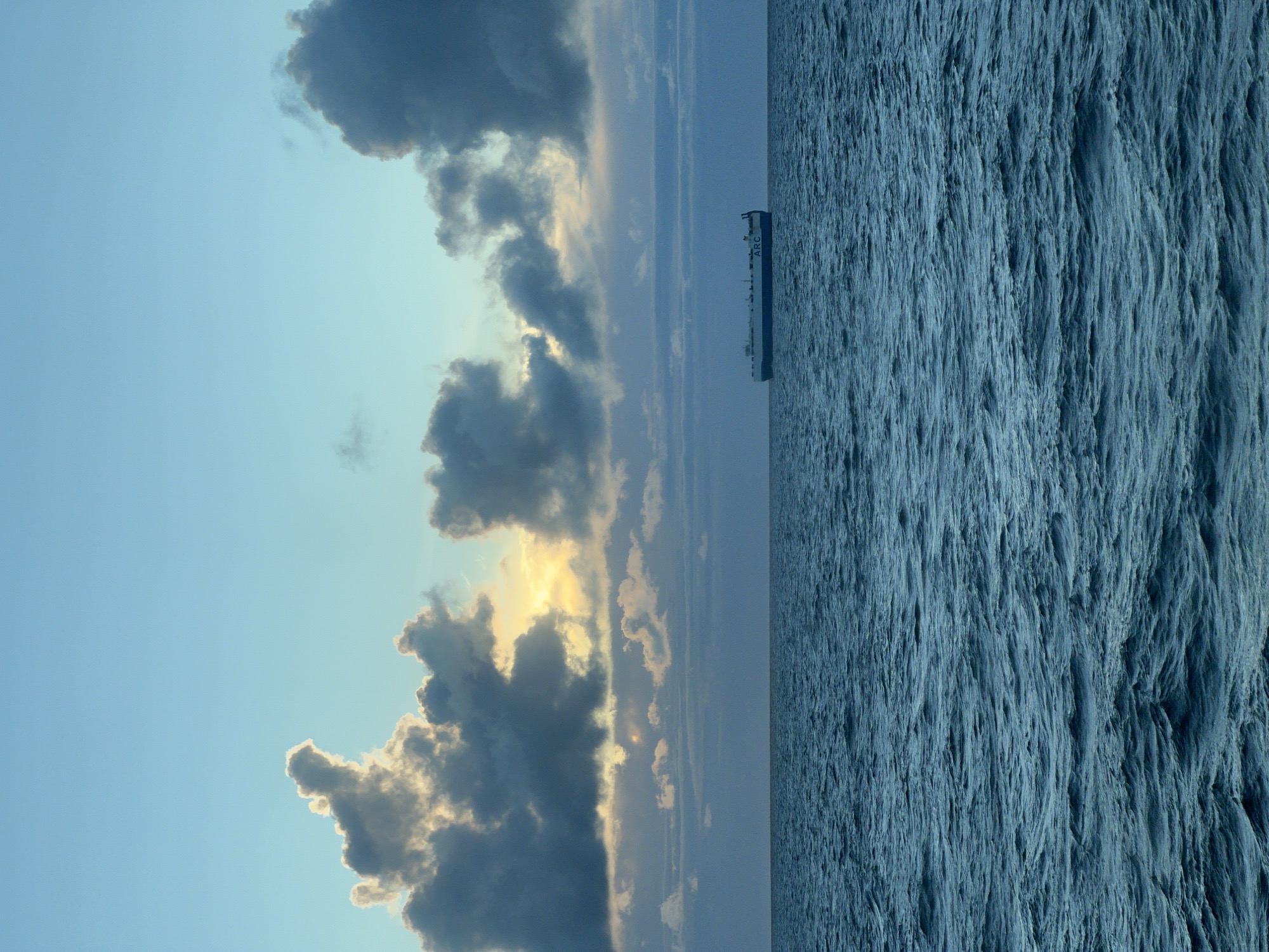 An ARC car carrier ship sails on a vast, choppy blue sea under a pale blue sky. Dark, dramatic clouds dominate the upper portion of the image, with a break in the clouds revealing a soft, golden sunset glow. The ship is positioned in the middle-right of the frame, appearing small against the immense expanse of the ocean and sky. The water is a deep, rich blue with visible waves and whitecaps, suggesting a moderately windy condition. The horizon line is clearly defined, separating the sea from the sky. There are no people visible in the image. The overall mood is serene yet dramatic, with the vastness of nature contrasting with the solitary presence of the ship. The location is unknown, with no specific city or country identified.