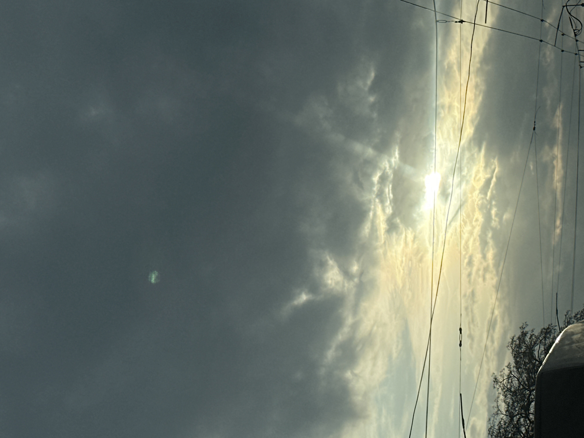 The image depicts a cloudy sky, likely taken during daylight hours. The sun is visible through a break in the clouds, casting a bright glow. Power lines are strung across the upper right portion of the frame. Part of a tree and a vehicle are visible on the lower right. The location in Kyiv, Ukraine, is not discernible from the image itself.