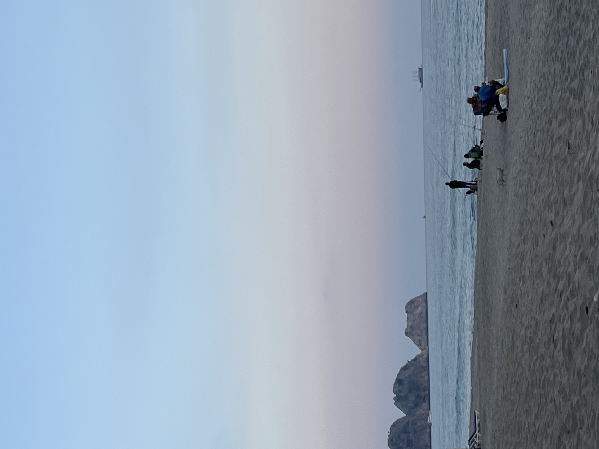 The image displays a serene coastal scene at what appears to be either dawn or dusk, characteristic of a beach in Alanya, Turkey. The sandy shoreline occupies the foreground and lower portion of the image, meeting the calm, gently rippling waters of the sea.
Several individuals are present on the beach, engaged in leisure activities. To the right, a small group is seated on beach chairs, with at least two individuals holding fishing rods extending into the water. Visible among them are people in a blue top and a green top. A light blue mat or board lies on the sand near this group. Further to the left, closer to the water's edge, a lone figure stands, also holding a fishing rod, with a small, light-colored dog standing nearby.
The sea stretches horizontally across the mid-ground. In the mid-distance, several dark, rugged rocky outcrops or small islands emerge from the water, adding visual interest to the landscape. On the far horizon, a large ship, possibly a cargo or cruise vessel, is faintly discernible, suggesting maritime activity.
The expansive sky above is clear and presents a subtle color gradient. It transitions from a pale, light blue in the upper left to a softer, hazy grey with hints of pink and purple towards the right and the horizon line. This soft lighting and color palette strongly suggest the scene is captured during the peaceful hours of twilight. The overall atmosphere conveyed is one of tranquility and quiet relaxation. There is no visible text in the image.