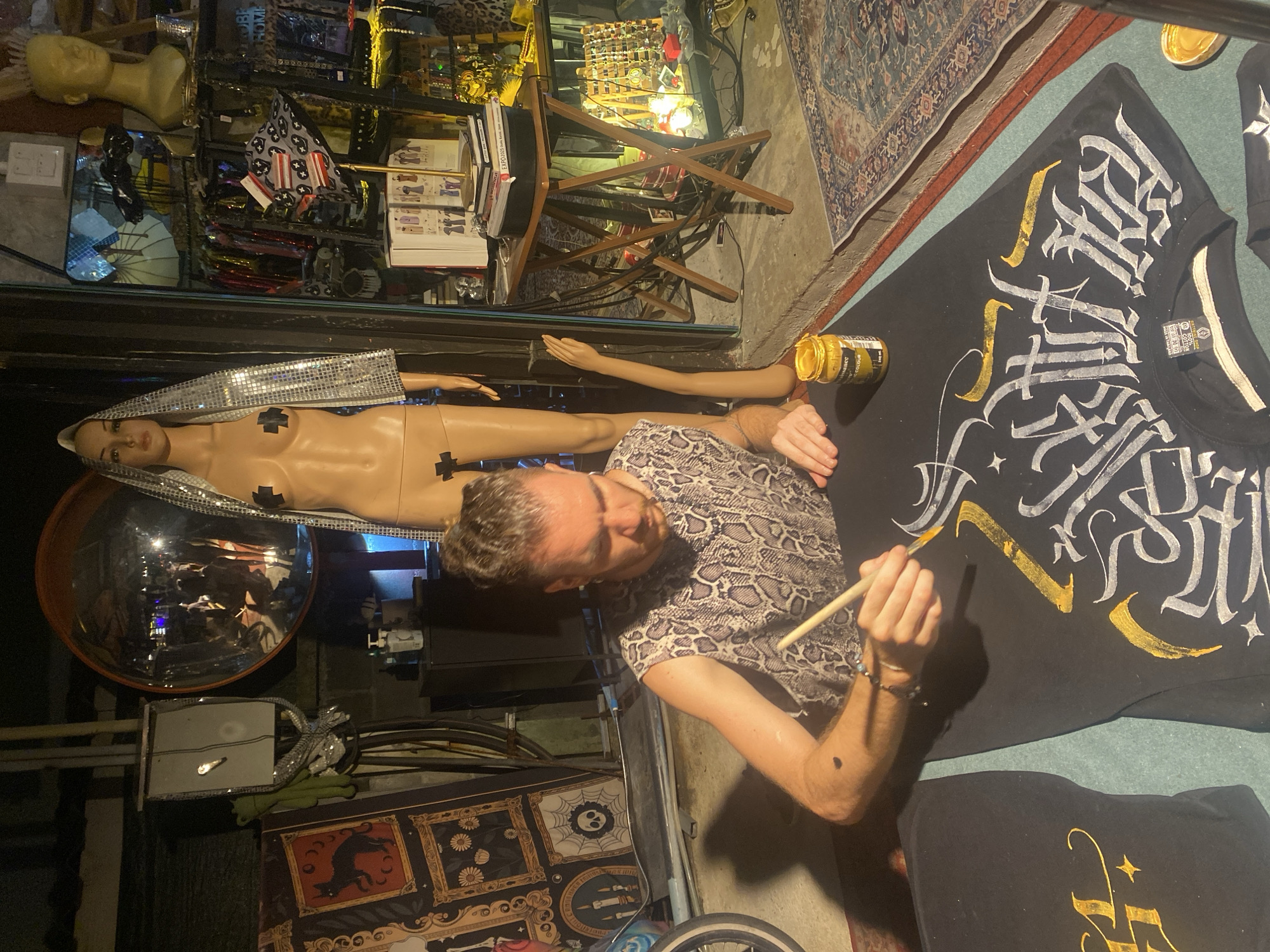 The image shows a man painting a black t-shirt with gold and white gothic-style lettering. He is using a wooden-handled brush and a jar of gold paint is visible nearby. The t-shirt is lying on a flat surface, possibly the ground, and another similar t-shirt is partially visible at the bottom.
The background includes a mannequin with a silver head covering, lying on its back behind a glass pane. A mannequin arm is extending from behind the glass toward the painter. Further back, there are shelves displaying various items, including other mannequins, flags, books, and lighting fixtures. There is also a round convex mirror reflecting the scene.
The location is suggested to be Baan Nai Suan, Thailand based on the presence of items and general aesthetic, though this is circumstantial. The surroundings are somewhat cluttered and artistic, indicating a creative or retail space.
