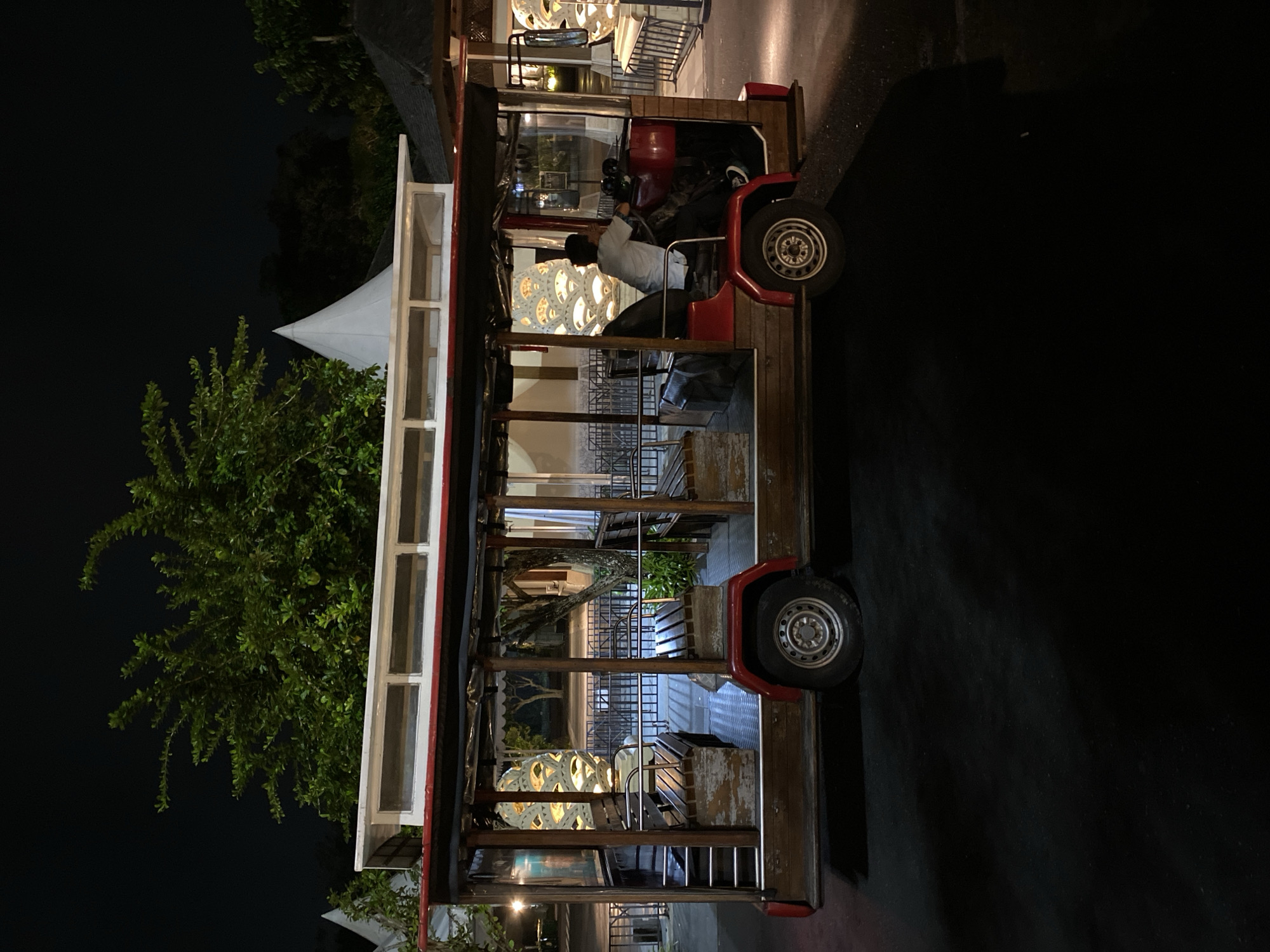 An open-air, wooden trolley with red trim is parked on asphalt at night. A person sits at the driver's seat, facing forward. The trolley is empty except for the driver. Behind the trolley, trees and a white tent-like structure are visible. The scene appears to be a resort or tourist area in Kuta Selatan, Indonesia, given the tropical foliage and resort-style architecture. The trolley is likely used for transporting guests around the property.
