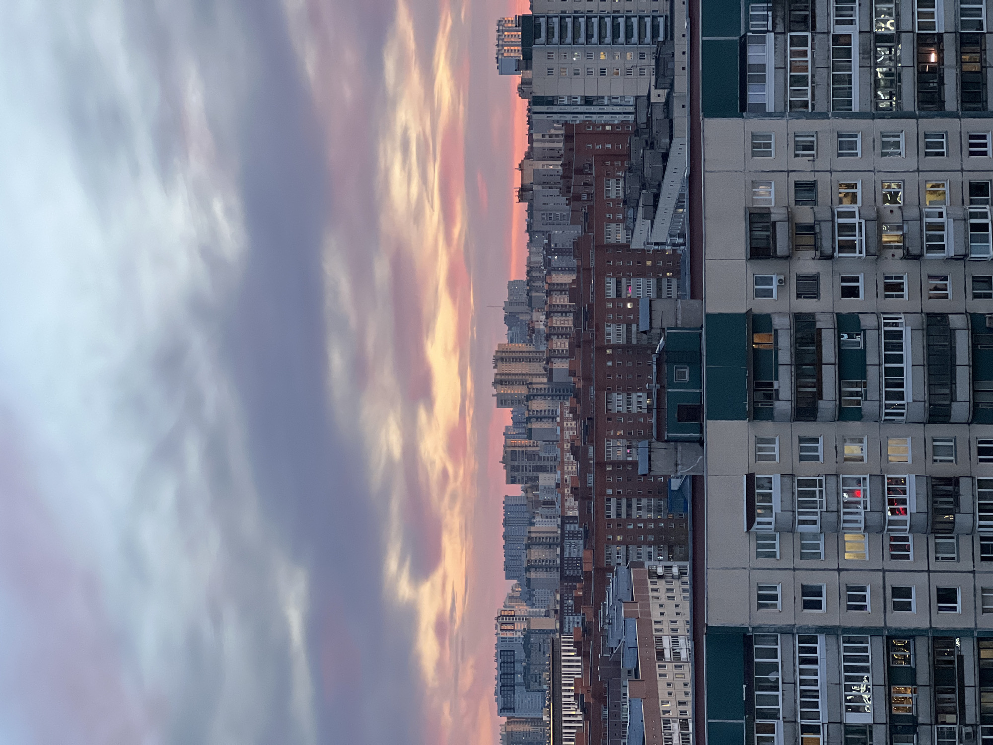 The image depicts a cityscape at twilight. On the right side are buildings with rectangular windows, some illuminated with light, suggesting interior activity. A large grey apartment building with dark green accents is in the immediate foreground on the right. Beyond the grey building is a mix of structures, including some constructed of brick, forming an urban skyline. Above the buildings on the left is a sky with a mix of purple and orange hues, indicating either sunrise or sunset. The scene is reminiscent of Soviet-era architecture, which is common in many Eastern European cities, particularly in Russia. The style and density of buildings suggest a location in a densely populated area like the outskirts of Saint Petersburg, Russia, where similar urban landscapes can be found. No people are directly visible in the image.