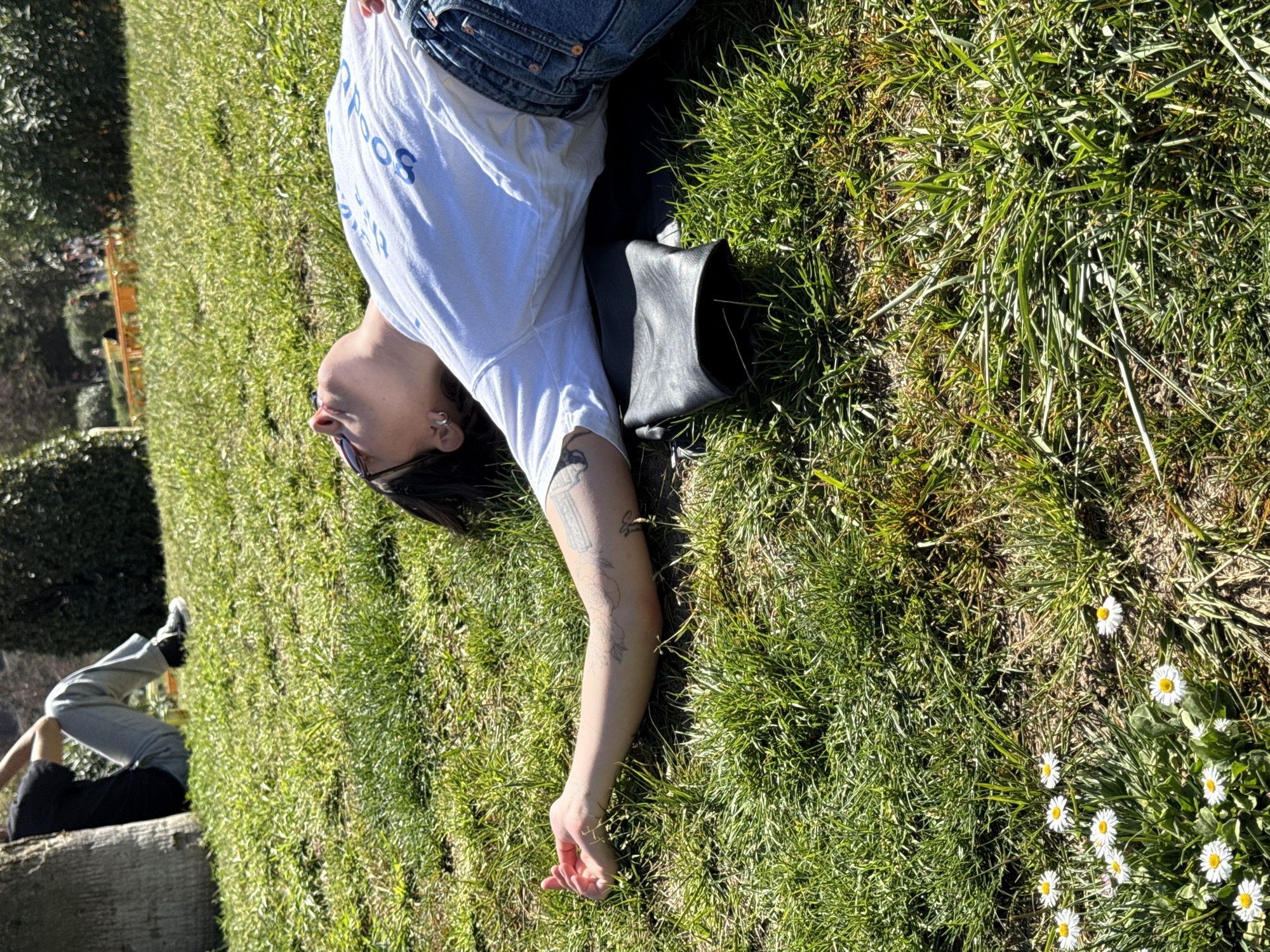 A person wearing jeans and a white t-shirt is lying on green grass in Barcelona, Spain. The person has dark hair, an earring, and a tattoo on their left arm. A black purse is next to them. Another person in light-colored pants and dark shoes is sitting on a concrete ledge in the background. There are daisies growing in the grass near the first person.