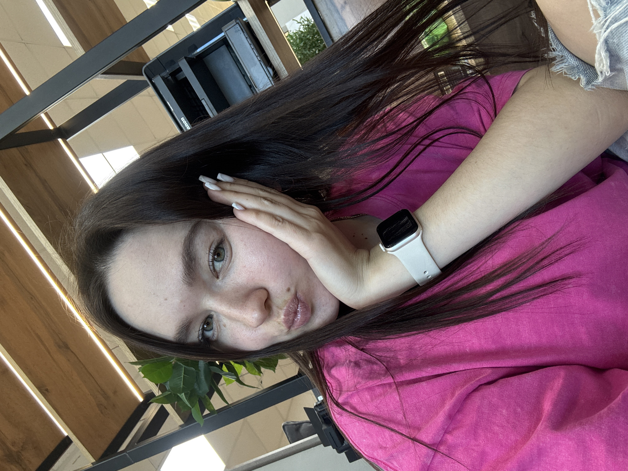 A woman with long, dark brown hair and green eyes is posing for a selfie. She is wearing a bright pink t-shirt and an Apple Watch with a white band on her left wrist. Her left hand is resting on her cheek. The background includes wooden ceiling slats, a black metal structure, and a plant. It is not possible to identify the precise location within the city of Chaikovsky, Russia based on the visual data provided.