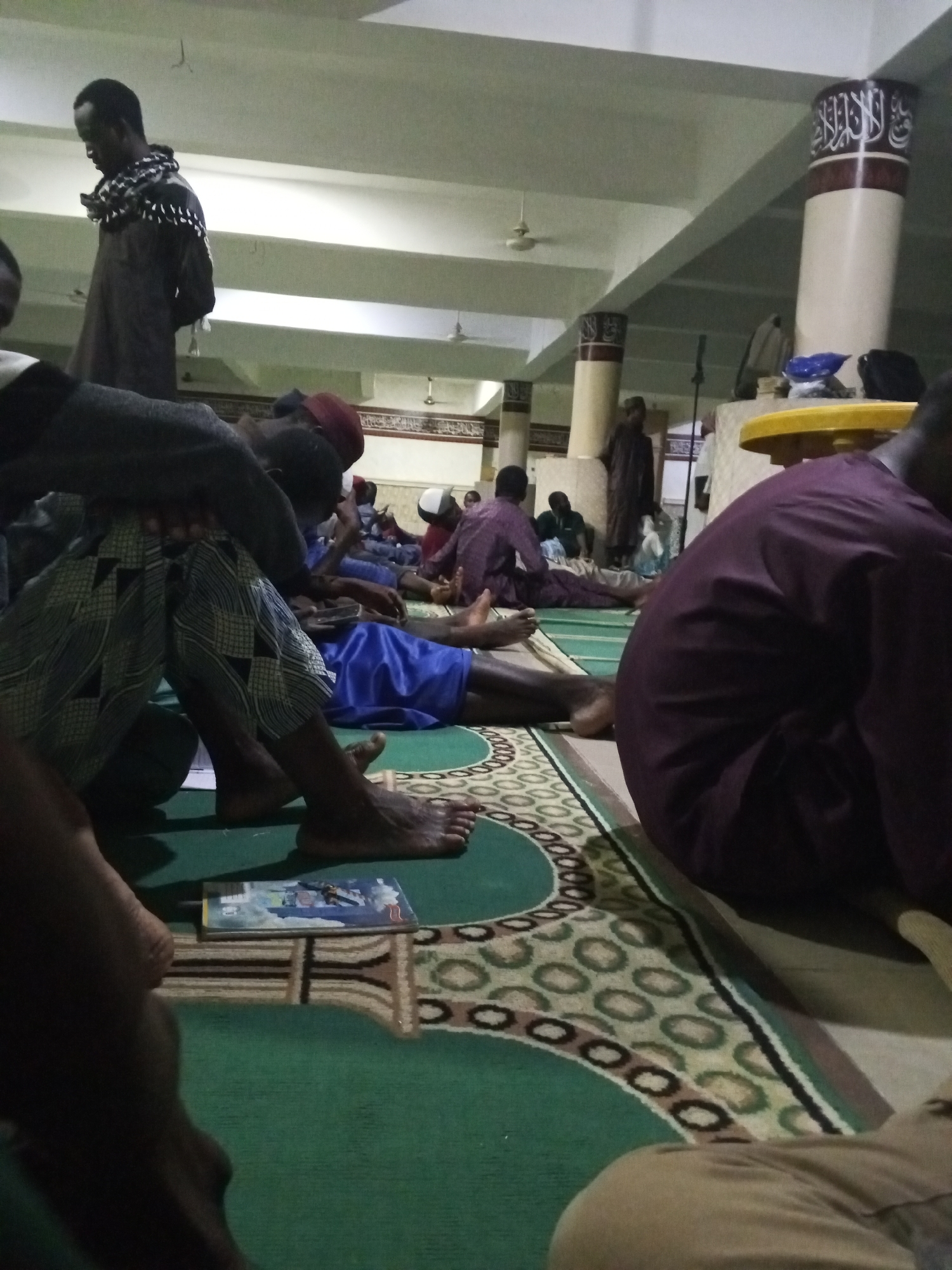 The scene appears to be inside a mosque in an unknown city in Nigeria. Several men are present, some sitting or reclining on the carpeted floor. The carpet is green with patterned borders. One man is standing, wearing a dark robe and a patterned scarf. There are pillars with Arabic script decorations. A book is lying on the carpet near a man's bare feet. A yellow table is visible in the background, and other individuals are dispersed throughout the space. The overall lighting appears somewhat dim.