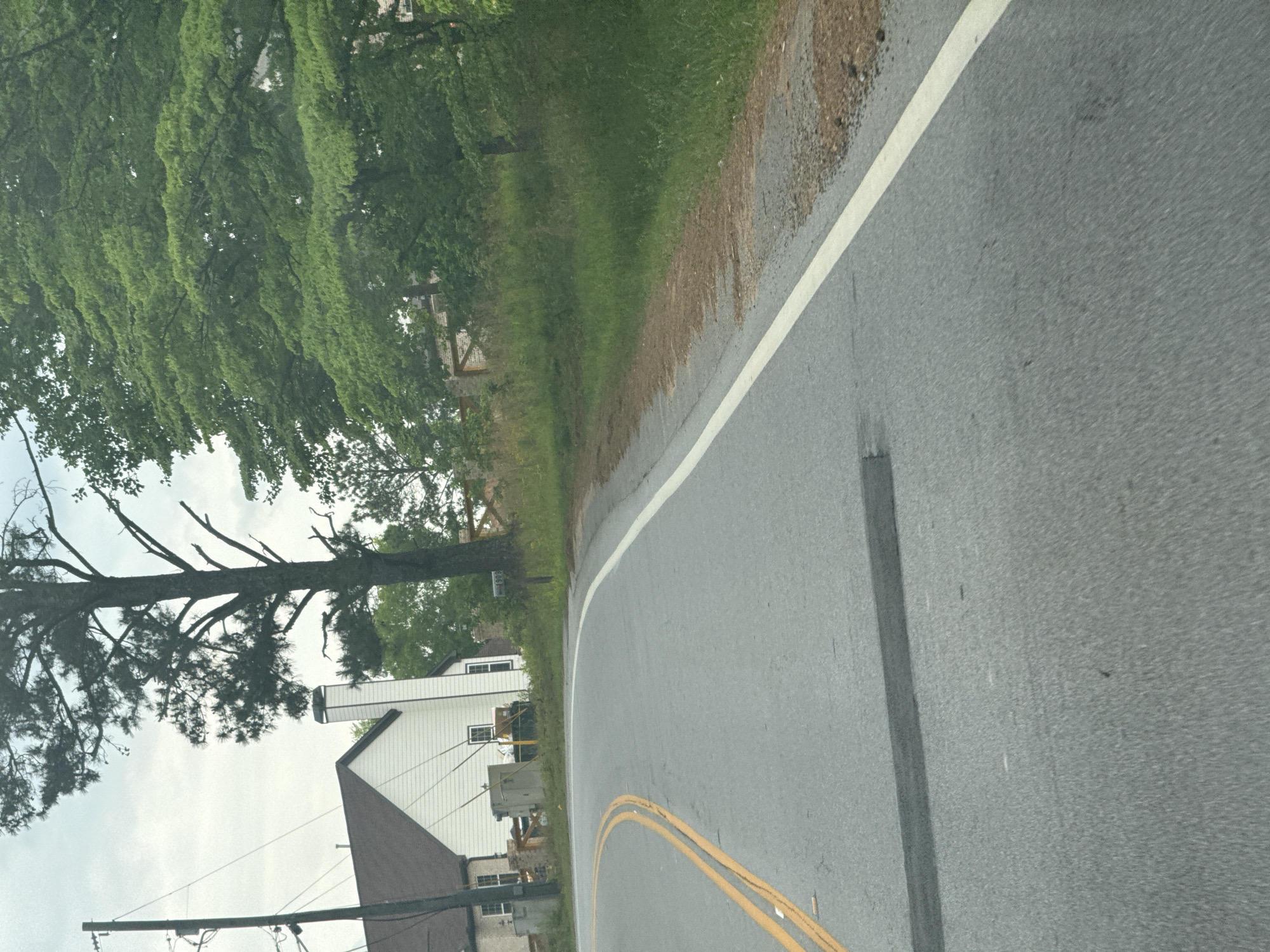 The image shows a view from a vehicle traveling on a road in Mountain Park, United States of America. The road has double yellow lines in the center and a white line on the right edge. To the left of the road, there's a grassy area with trees and a house with a white exterior and a brown roof. Utility poles and wires are also visible. The sky is overcast.