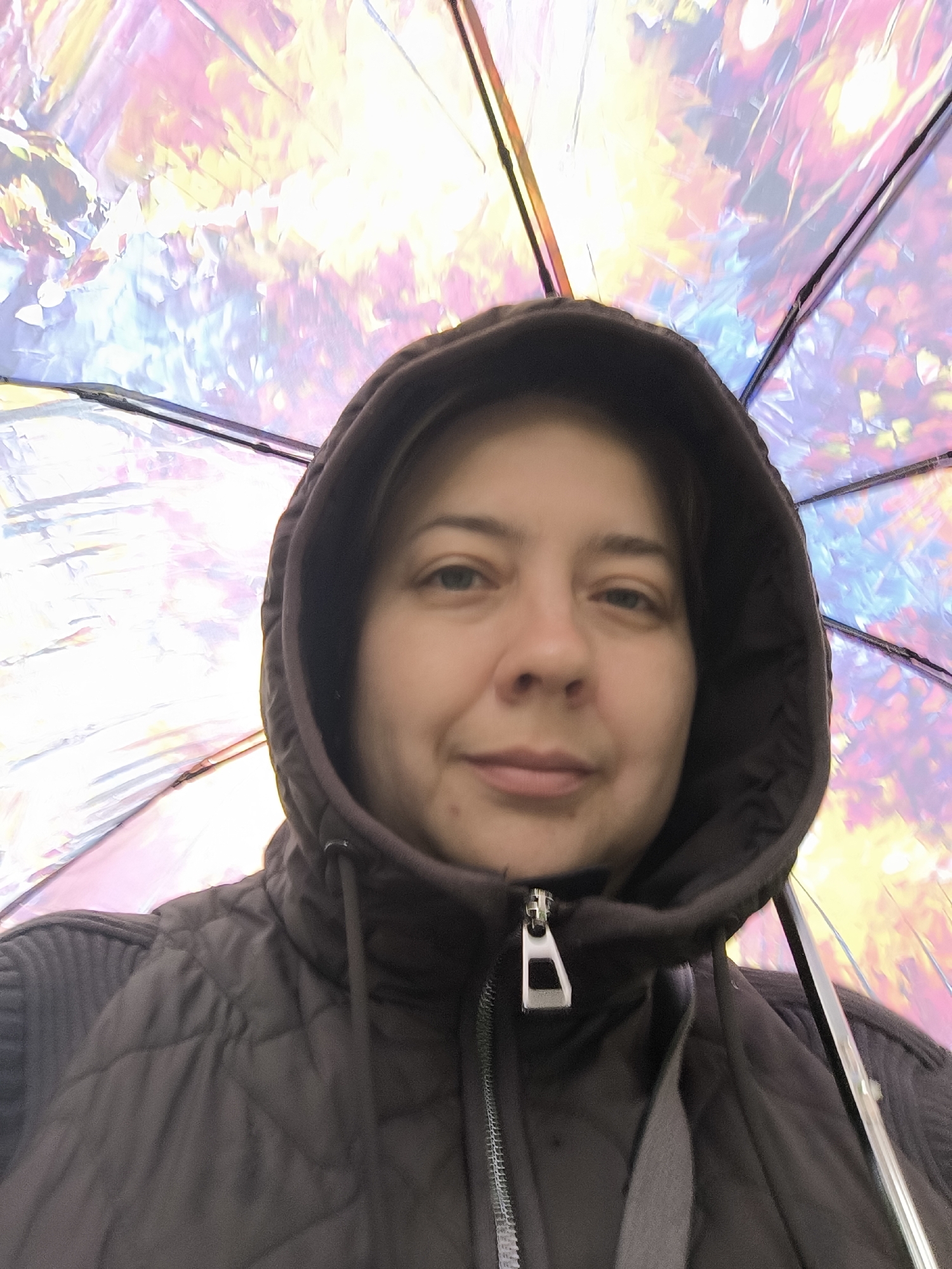 A woman is centered in the frame, standing underneath a multicolored umbrella. She is wearing a dark hooded jacket and is looking directly at the camera. The umbrella above her is iridescent with hues of pink, yellow, blue, and white. The background is not discernible, providing no clear indication of the location in the city of Unknown, Unknown.