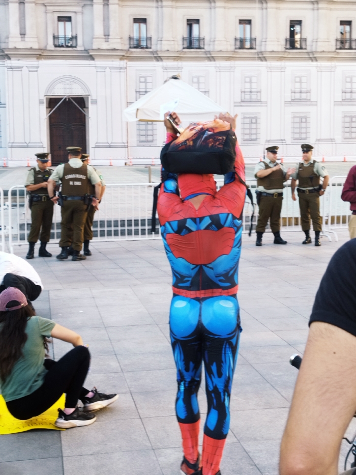 A person dressed as Spider-Man stands in a public square with their back to the camera. They're wearing a backpack. In the background, police officers are standing and watching. To the left, a woman is sitting on the ground. The setting appears to be in front of a large building with many windows. The scene suggests a public event or gathering where people are interacting and being observed by law enforcement.