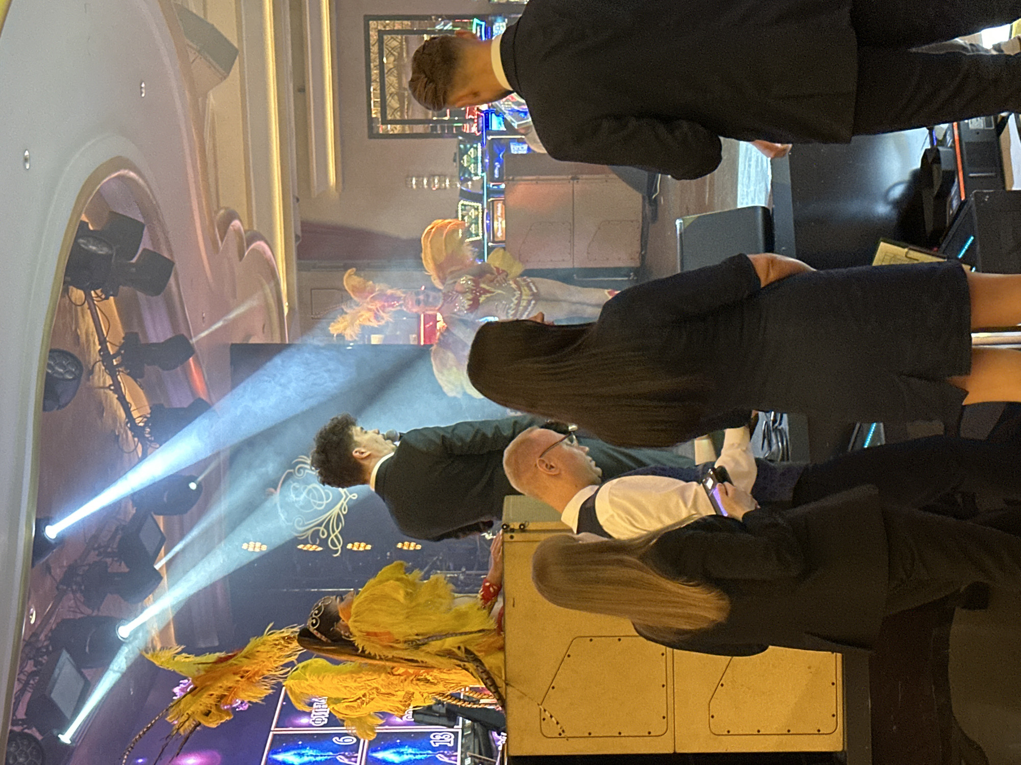 In Estosadok, Russia, a male performer in a black suit stands on a stage, holding a microphone, as bright stage lights illuminate the scene. To his left, a female performer in a yellow feathered costume, possibly a dancer, is positioned near a large yellow speaker cabinet. In the foreground, three individuals are visible: a woman with long brown hair wearing a black blazer and pants, looking down at a mobile device; a man wearing glasses and a white shirt with a dark vest, also looking downwards; and a woman with long dark hair wearing a dark blue dress, facing away from the camera. Behind the stage performers, slot machines are visible, suggesting the location might be a casino or entertainment venue. The overall environment appears to be an indoor event with a performance and audience present.