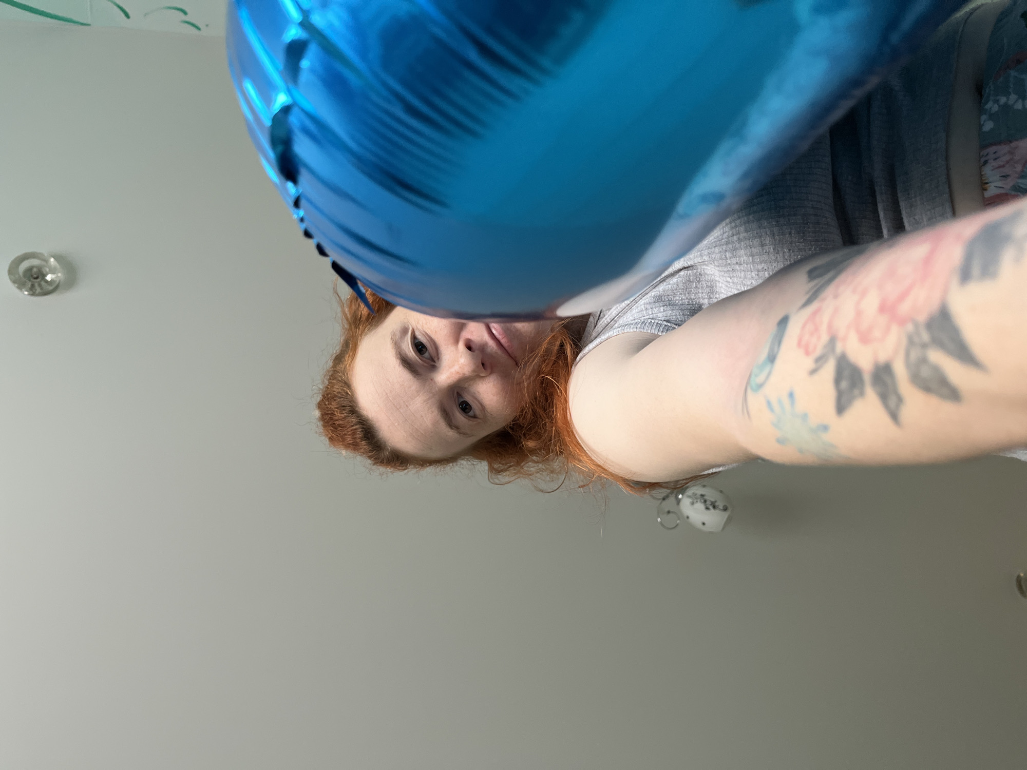 A woman with red hair and a tattoo on her arm is holding a blue balloon. The woman is looking down at the camera. The background is a gray wall with a light fixture and a door visible. The location is presumed to be an indoor space based on the interior design elements present in the image. It is not possible to confirm or deny that this image was taken in the city of Chaikovsky, Russia.