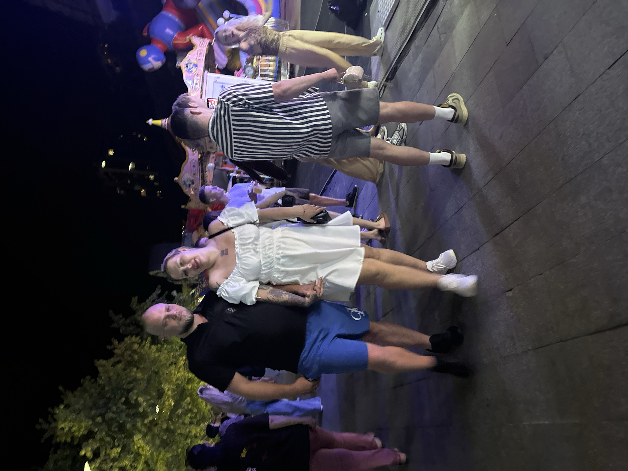 At night, a couple stands holding hands, facing the camera. The man, on the left, has a bald head and a black polo shirt. He wears blue shorts with a white logo. The woman, on the right, has blonde hair and wears a white off-the-shoulder dress. She has tattoos on her arm. They are at an outdoor fairground with rides and other people in the background. The image is taken at night, possibly in a city. The location is not Odesa, Ukraine.