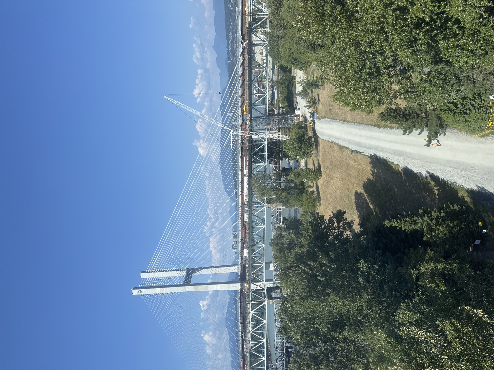 The image captures a sunny day in Surrey, Canada, with a focus on the Alex Fraser Bridge. The bridge, a prominent cable-stayed structure, is visible in its entirety, featuring its distinctive twin concrete towers and numerous suspension cables. Construction appears to be ongoing on the bridge's deck, with a large white crane positioned near the center, extending its arm over the structure.
Below the bridge, a wide, unpaved gravel road stretches into the foreground, winding through a park-like setting. The road is bordered by lush green trees on both sides, with patches of dry, golden grass visible in the background. There are a few small yellow construction vehicles and some construction workers in high-visibility vests on the road, suggesting ongoing work in the area. The sky is a clear, vibrant blue, with a few distant mountains visible on the horizon. The presence of the bridge and the general landscape suggest this location is in a semi-urban or suburban area with significant natural elements.