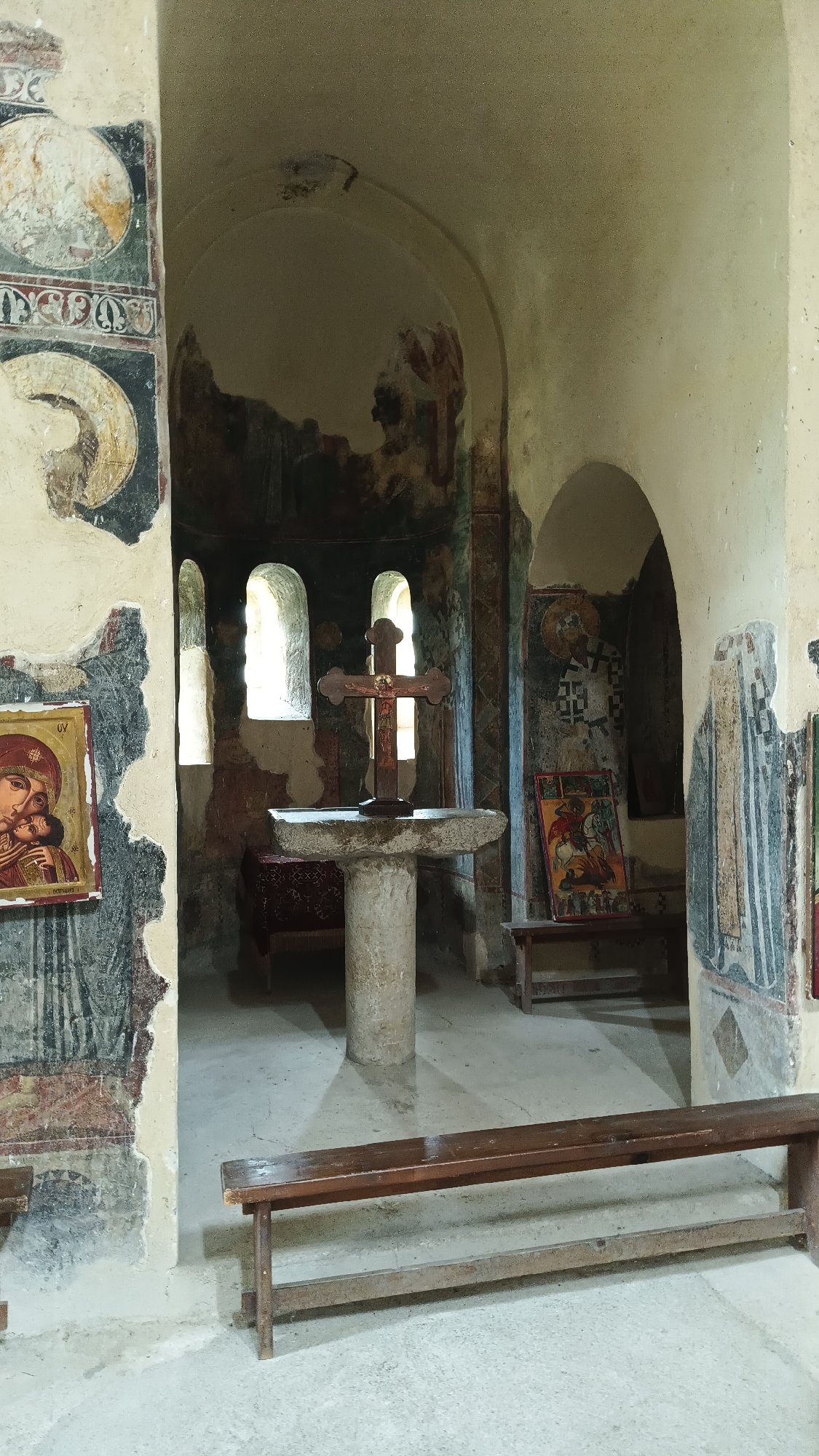 The image shows the interior of a church in an unknown city. The space is worn and has an aged look, with weathered walls displaying faded frescoes. In the center of the room is a stone altar with a wooden cross on top. Behind the altar, there are arched windows that let in natural light. Religious icons are placed on the walls, and wooden benches provide seating for visitors. The overall atmosphere is one of quiet reverence and historical significance.
