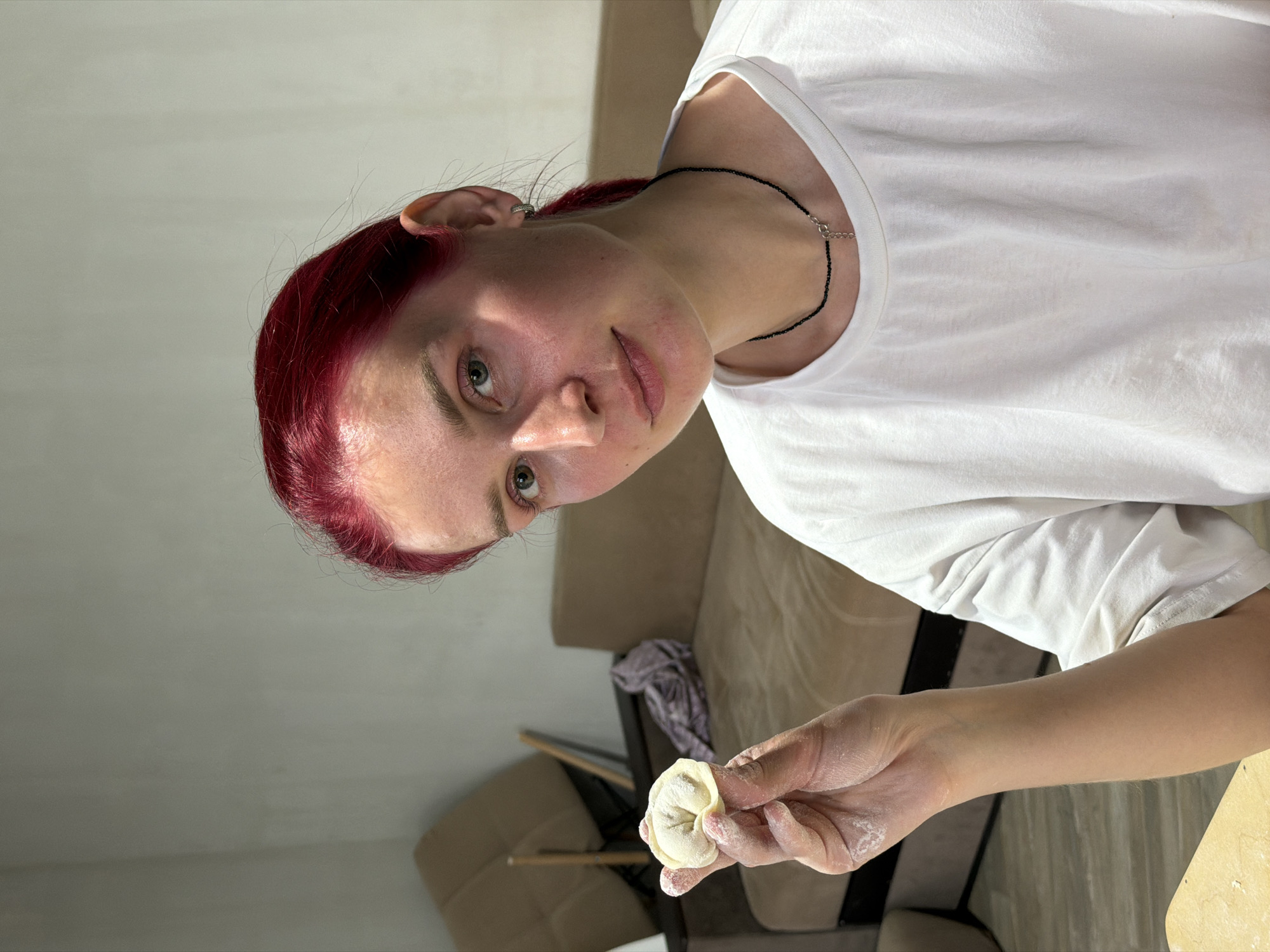 The image shows a person with red hair wearing a white t-shirt and a black necklace. They are holding a pelmen, a type of dumpling, in their flour-covered hand. The background includes a beige sofa and what appears to be part of a wooden chair. The photo was taken indoors, but the location in the city of Chaikovsky, Russia is not evident from the image.