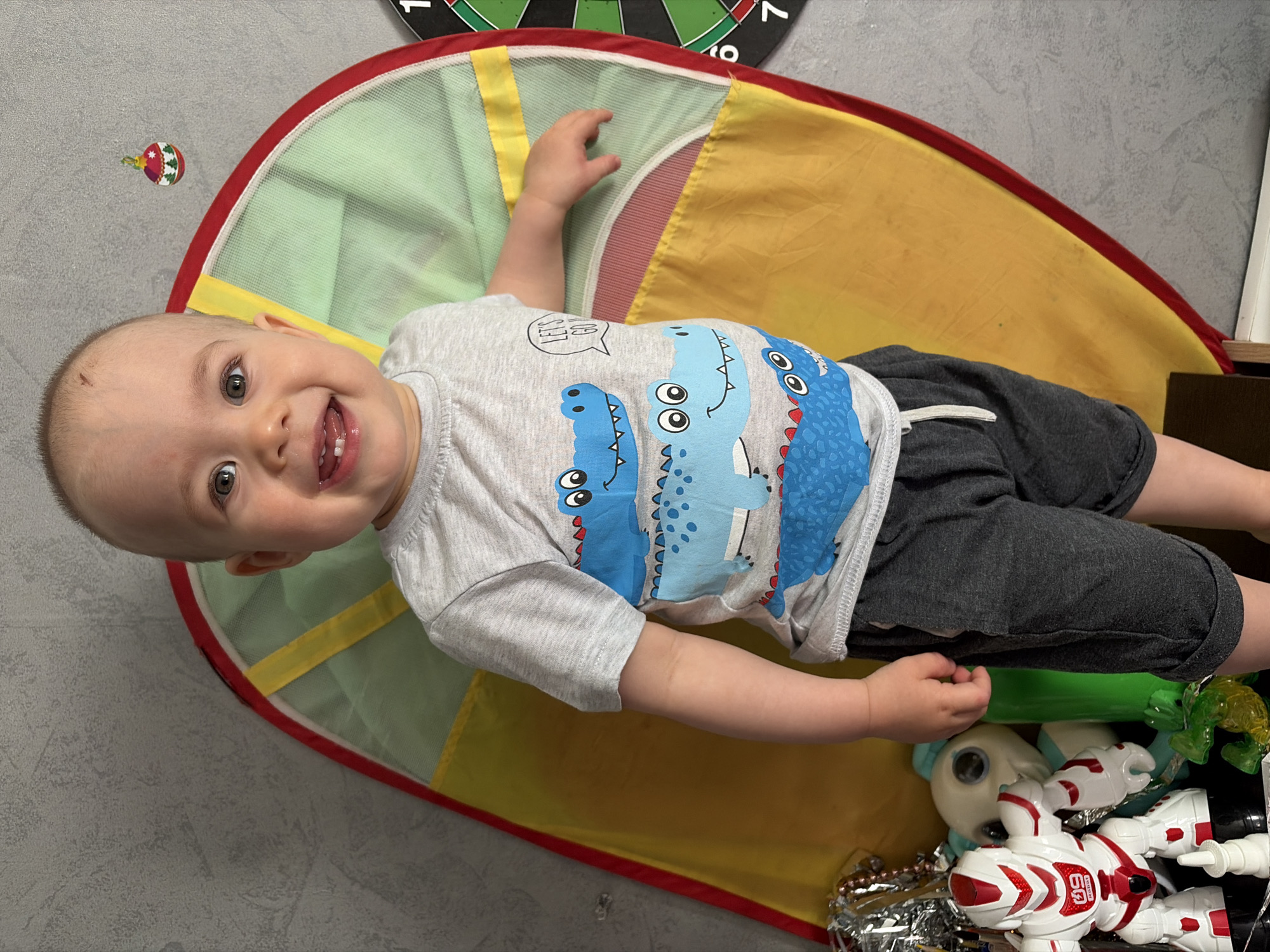 A young child is lying on a colorful play mat, smiling at the camera. The mat is predominantly green and yellow with a red border. The child is wearing a gray T-shirt with blue dinosaur prints and dark gray shorts. To the right of the child, there are toys, including a white and red robot. In the background, a dartboard is visible, partially cropped. The scene appears to be indoors, on a gray textured floor. Based on the location information, this image was likely taken in Chaikovsky, Russia.